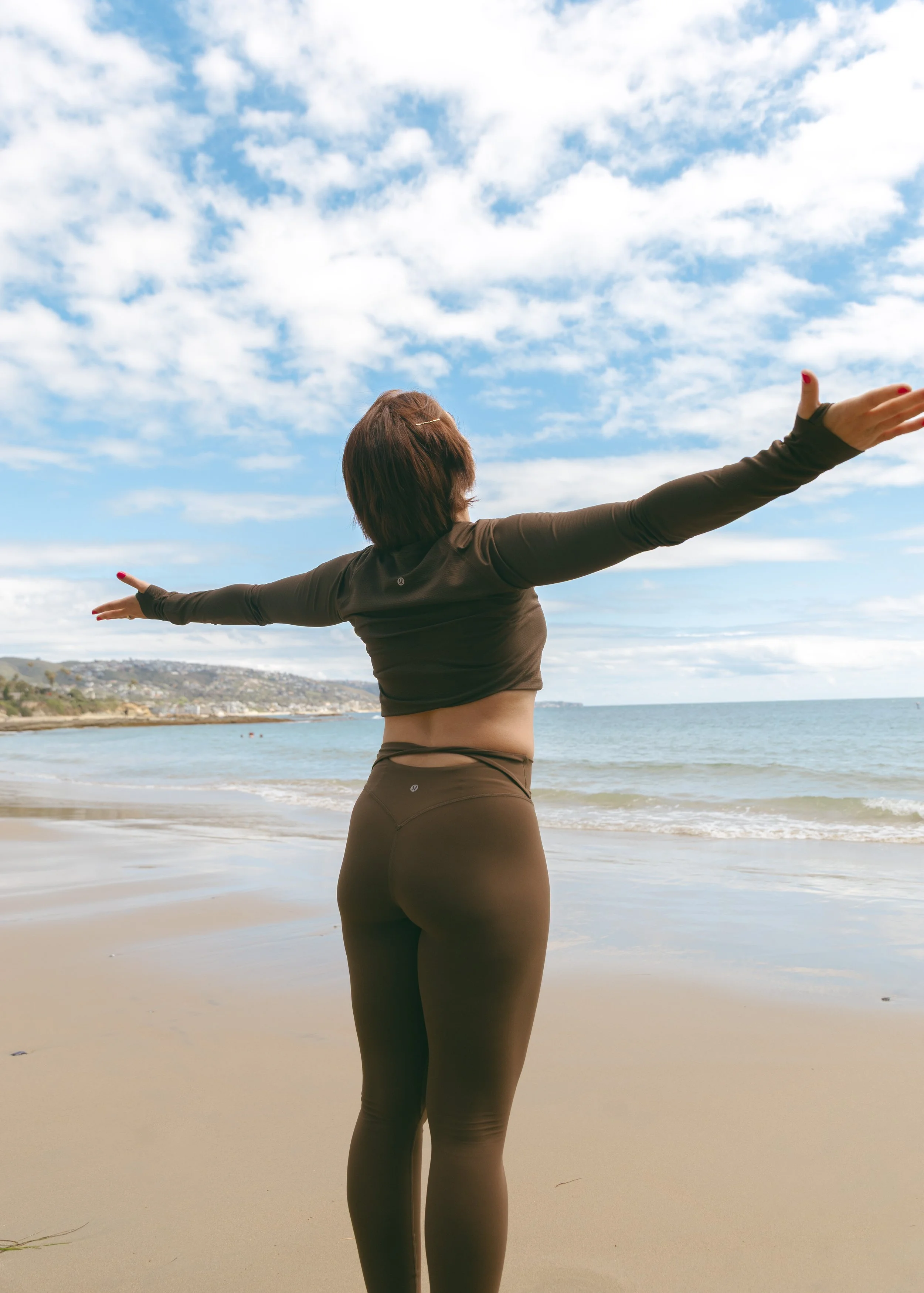 Photo of a woman with her back to the camera standing in front of the shoreline with her arms stretched out.