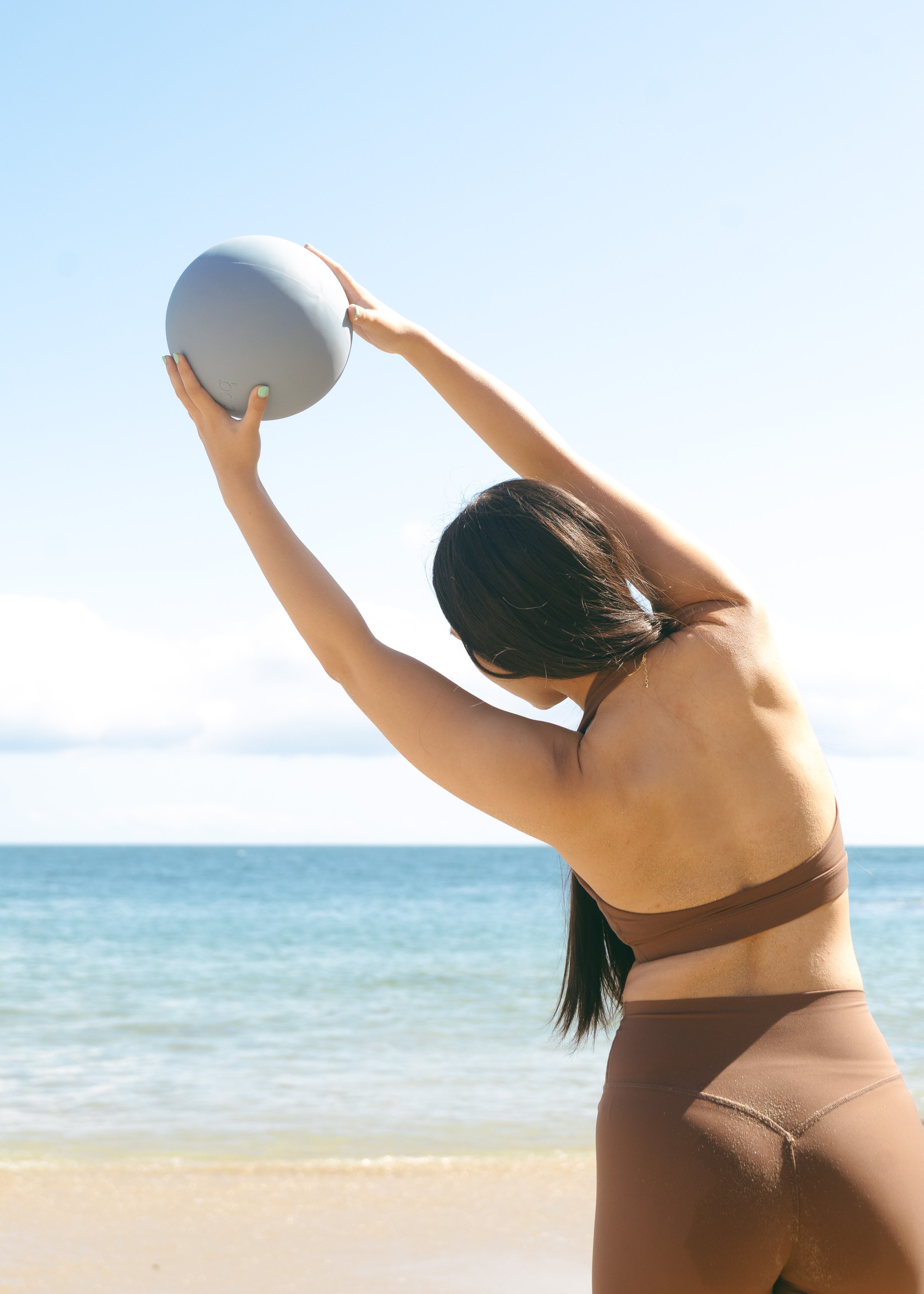 Photo of a girl in a matching yoga set with her back turned towards the camera standing in front of the shoreline holding a blue ball over her head.