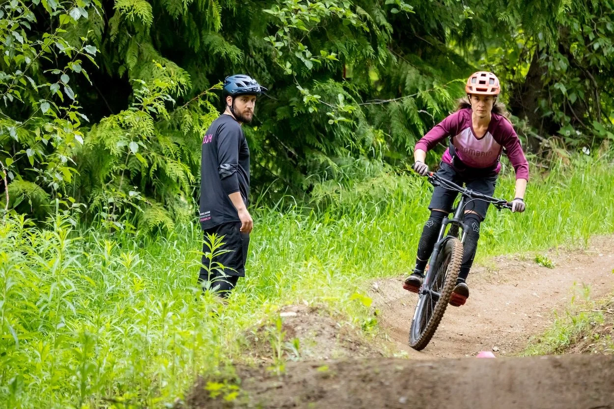 A woman mountain biking on a dirt trail with a man standing nearby in a green outdoor forest setting.