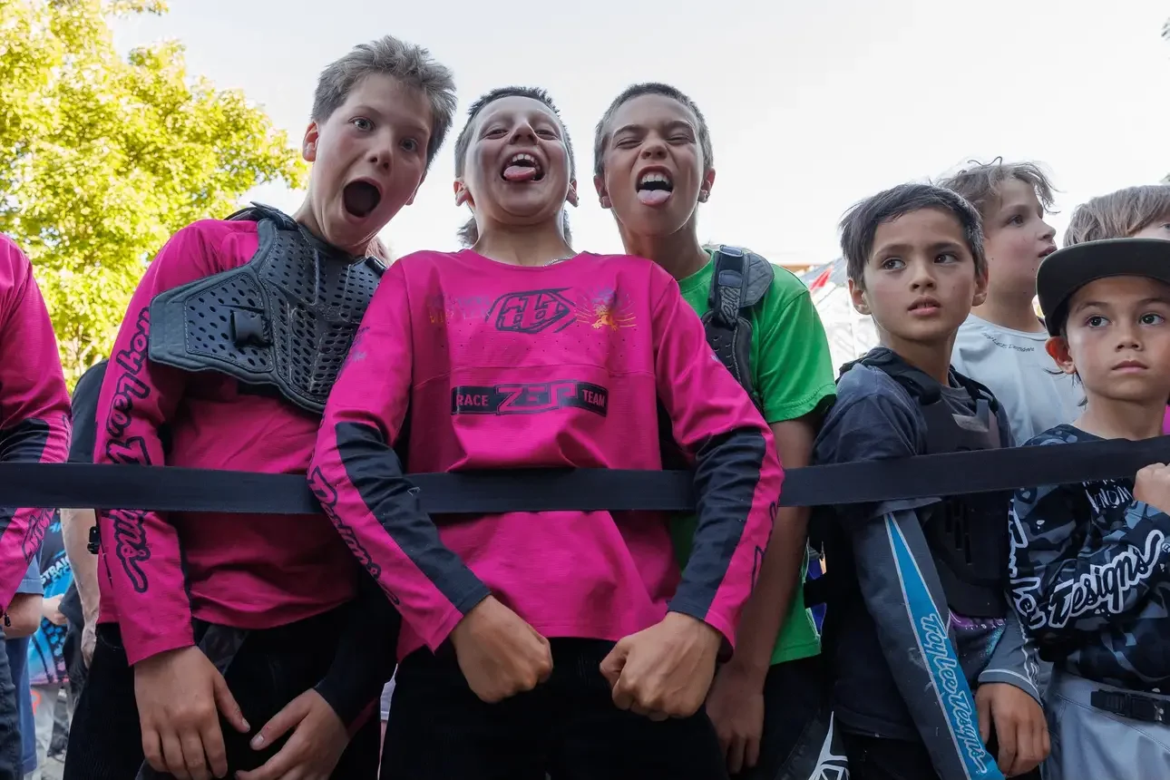 Group of children with excited and intense facial expressions, standing behind a black barrier, outdoors during daylight.