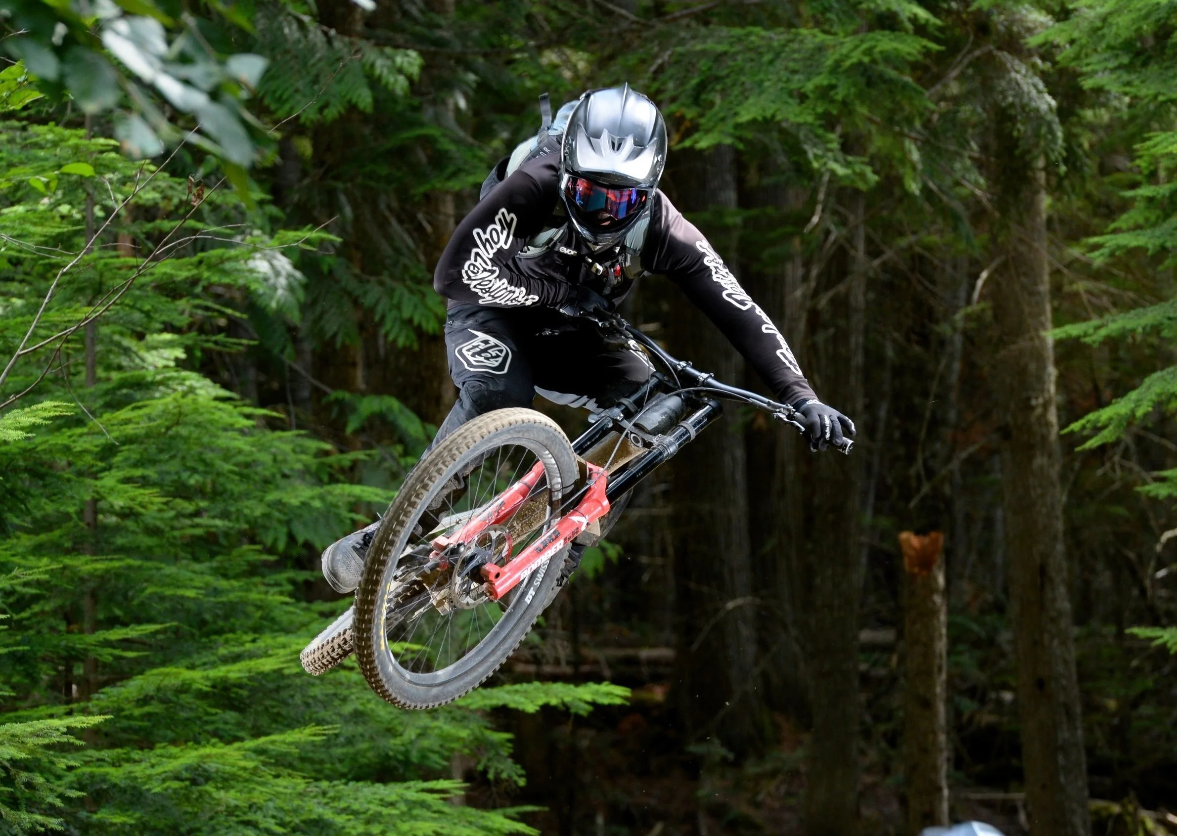 A mountain biker wearing a black helmet and black gear jumps off a trail in a forested area.