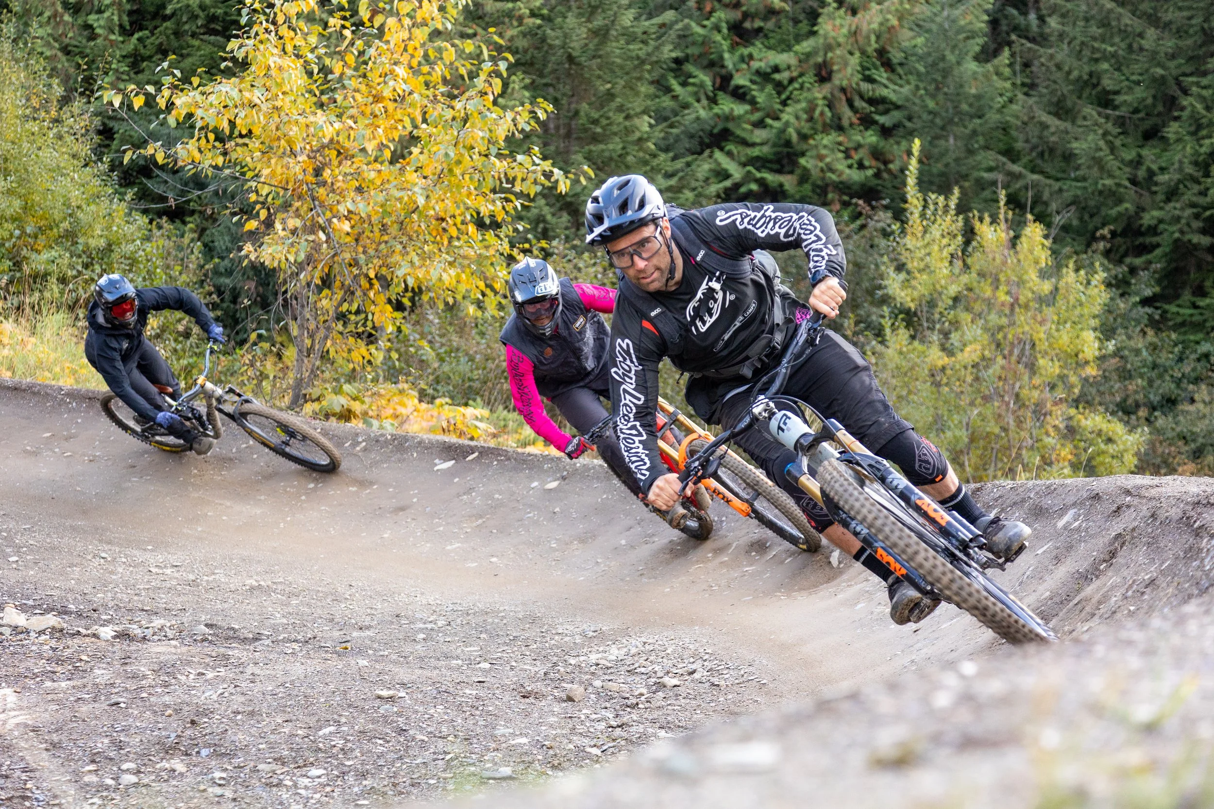 Three mountain bikers wearing helmets and protective gear riding downhill on a dirt trail surrounded by trees with autumn foliage.
