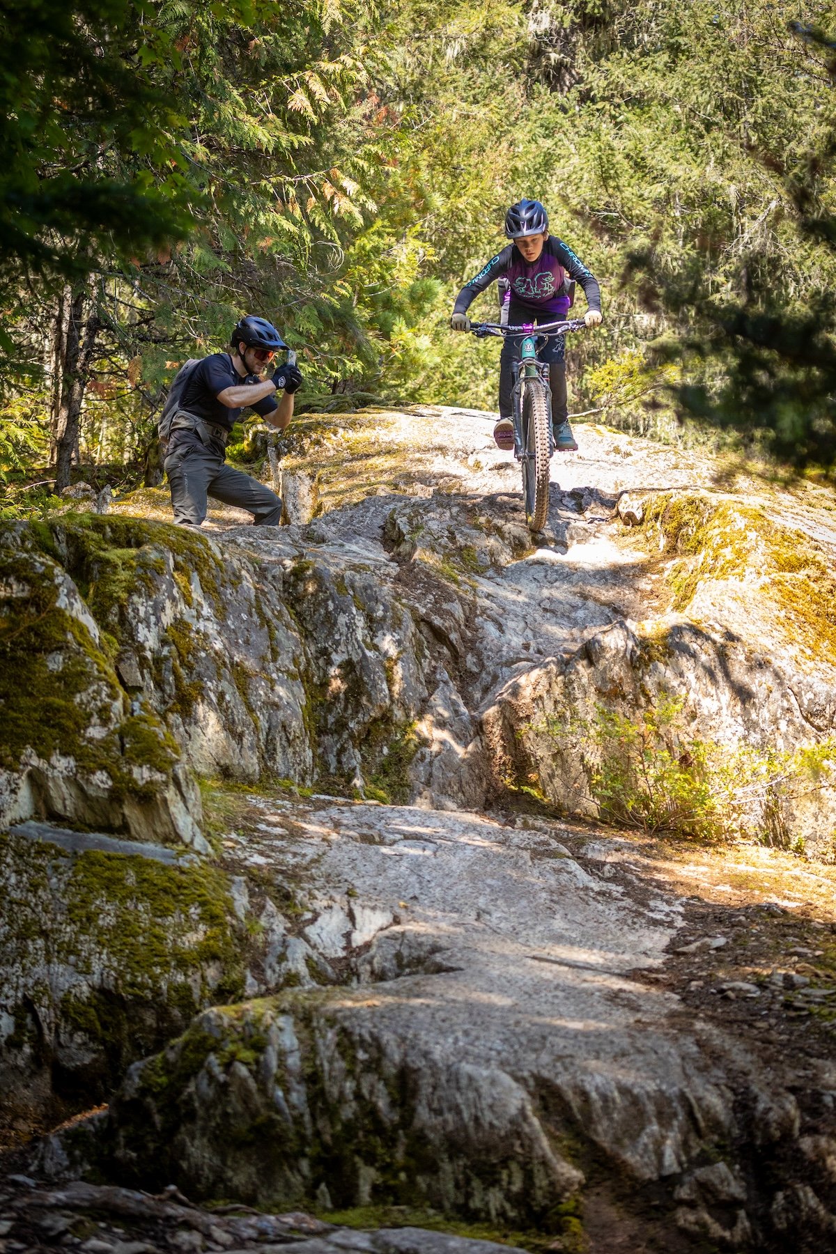A young girl is mountain biking down a rocky trail in a forest, with a photographer crouched nearby taking pictures.