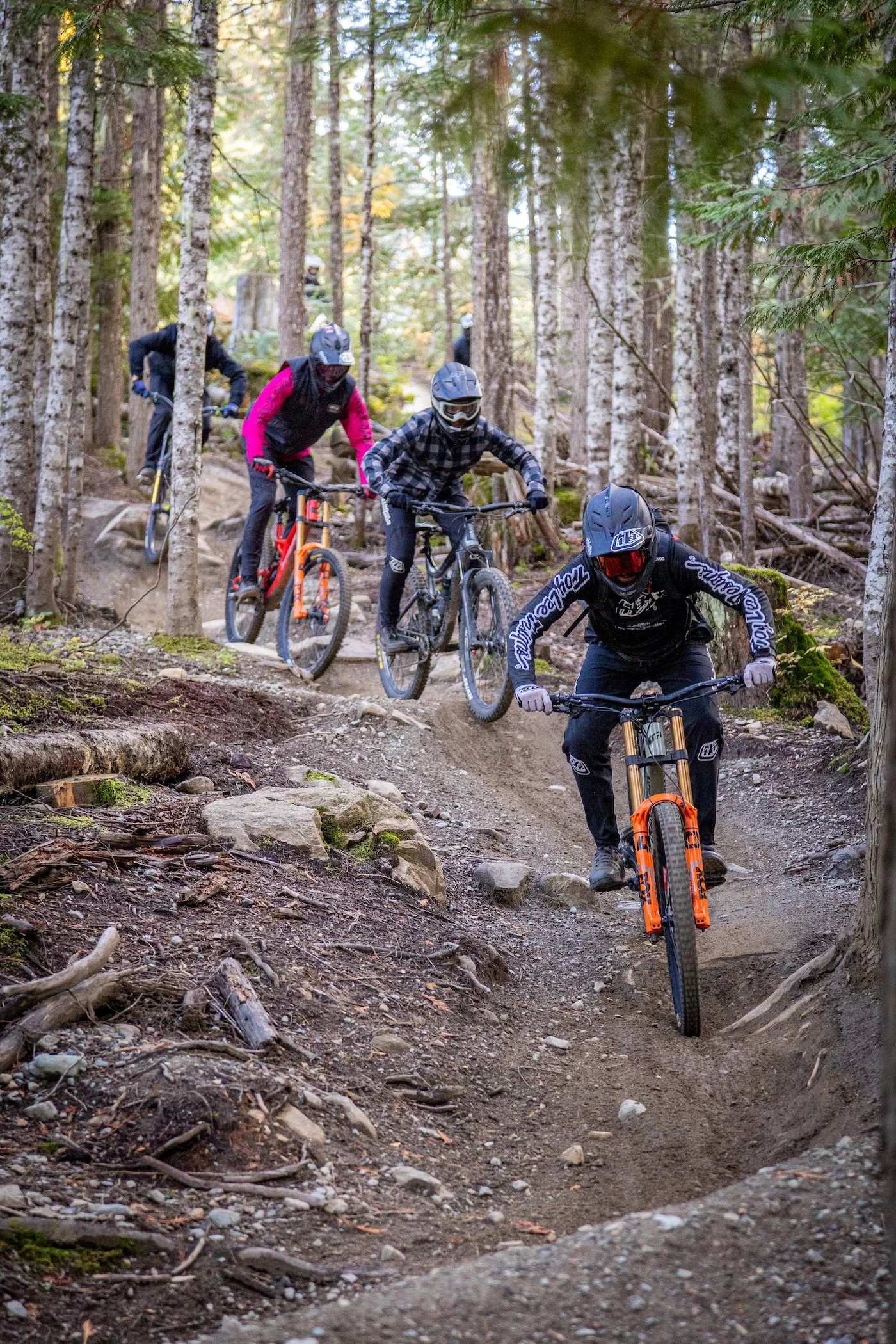 Group of mountain bikers riding down a trail in a forest, wearing helmets and protective gear.