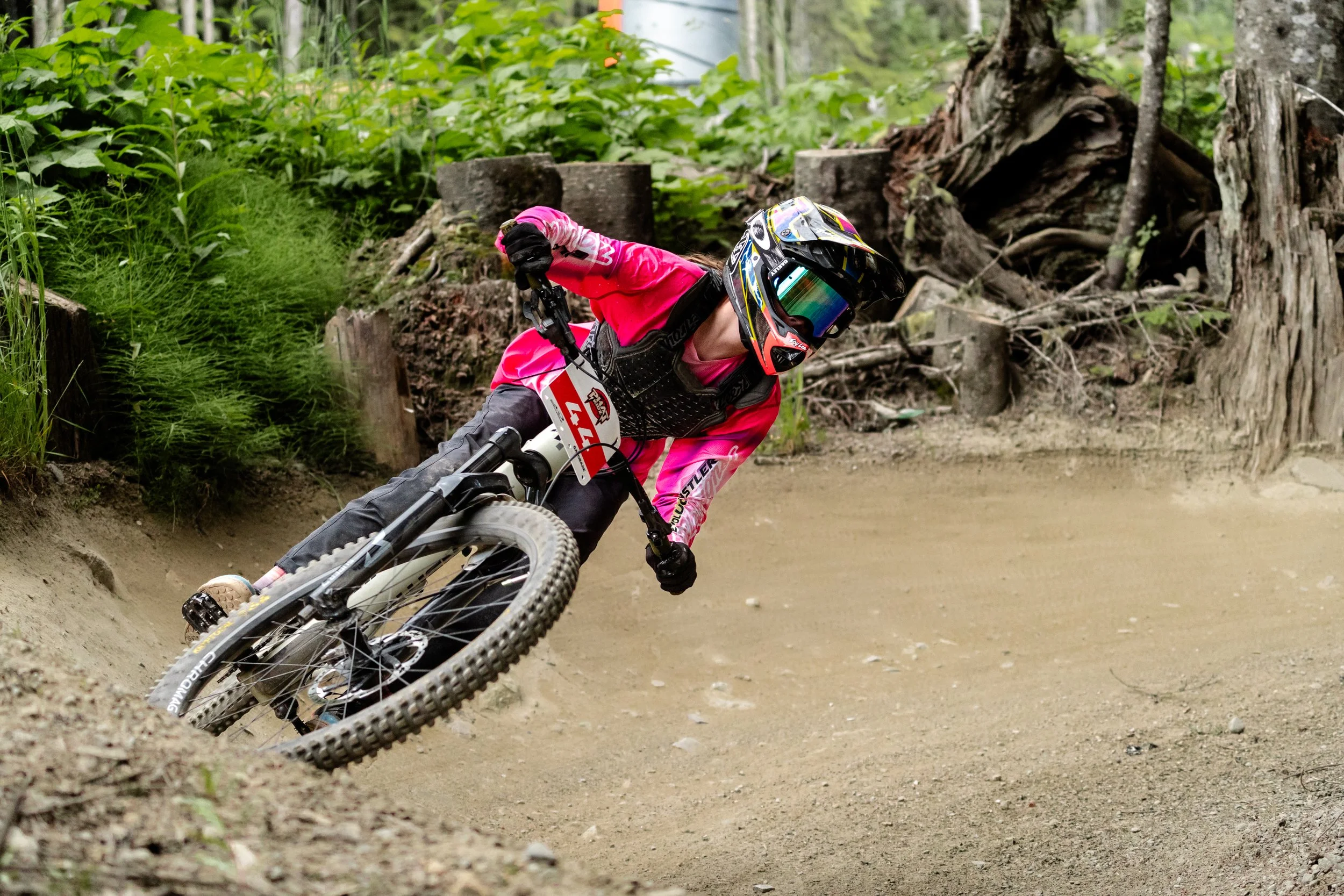 Mountain biker wearing a pink jersey and black helmet leaning into a turn on a dirt trail in a forest.