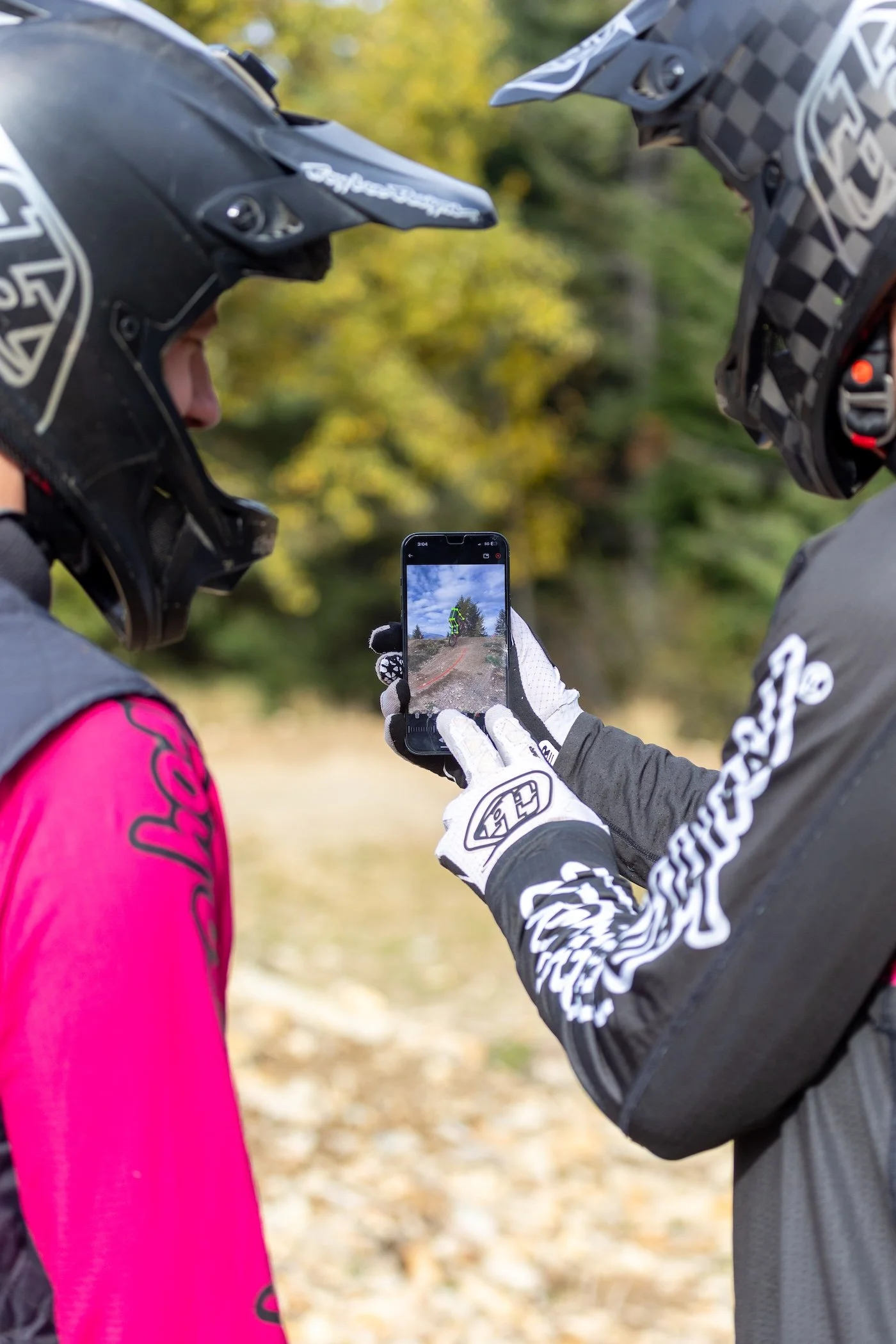 Two mountain bikers wearing helmets and gloves reviewing a trail map on a smartphone outdoors in a wooded area.