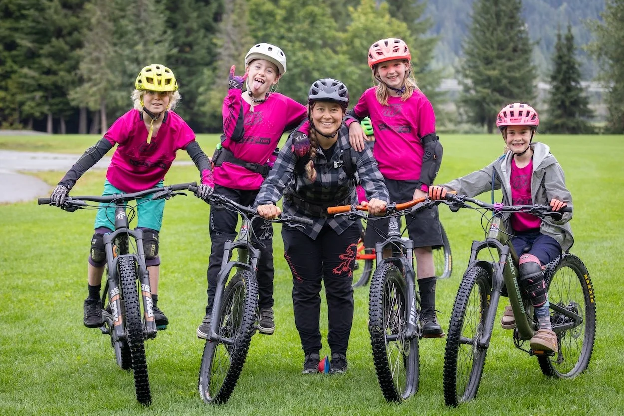 Group of four girls and one woman with bikes, wearing helmets and protective gear, smiling outdoors on grassy field with trees in background