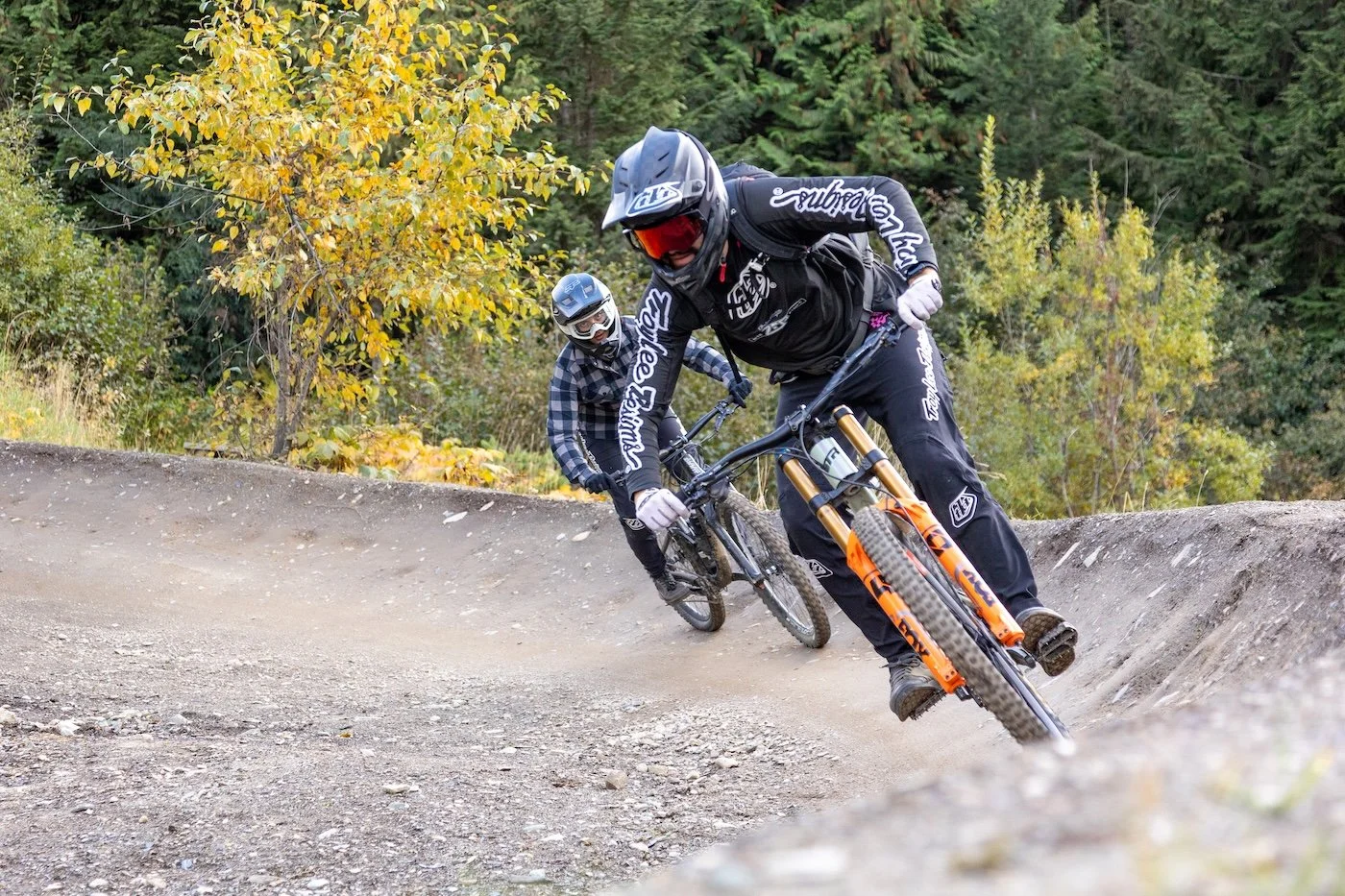 Two mountain bikers in protective gear riding downhill on a dirt trail in a forested area with trees and autumn foliage.
