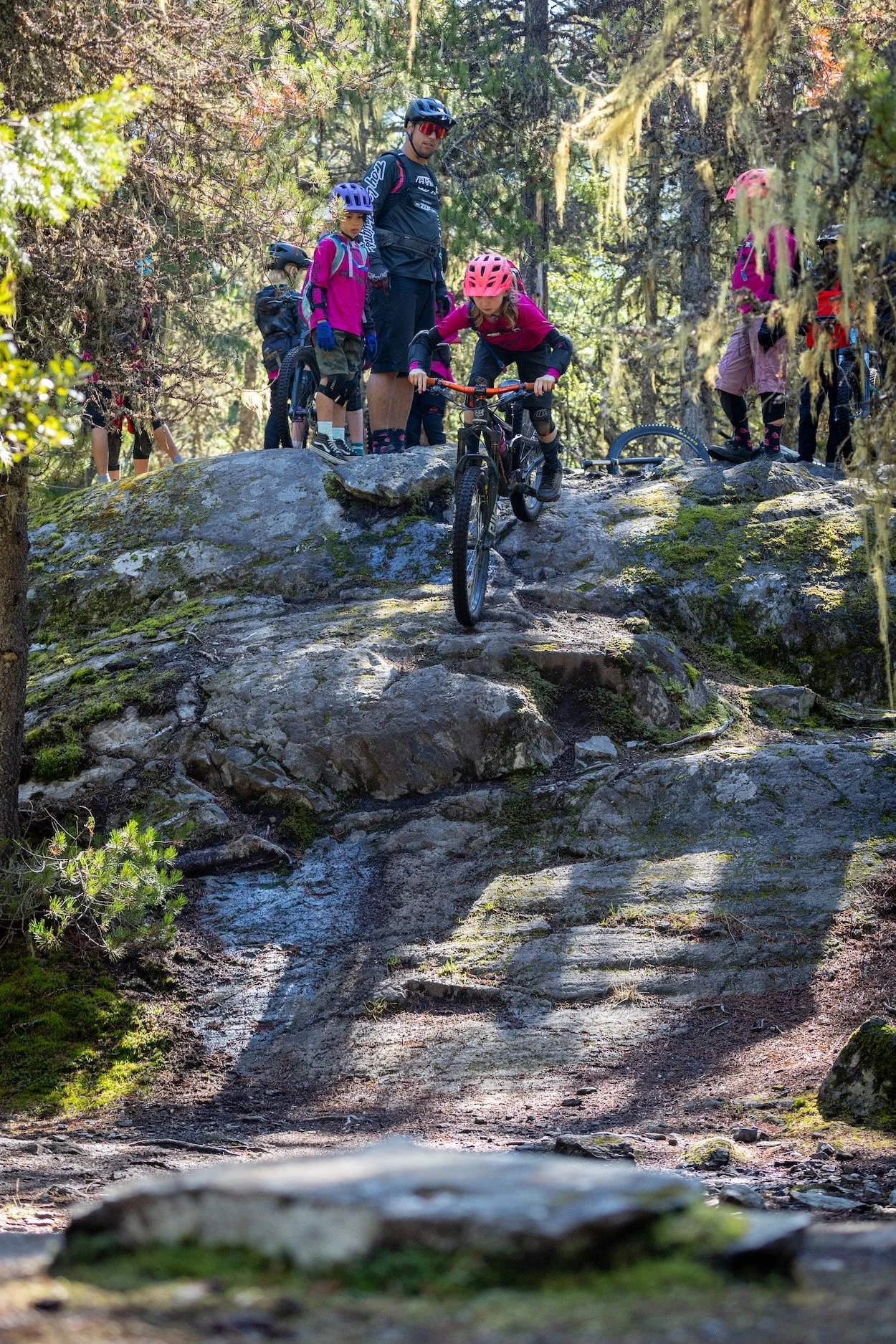Children and adults on a mountain biking trail in a forest with rocks, moss, and trees, some children wearing pink helmets and backpacks.