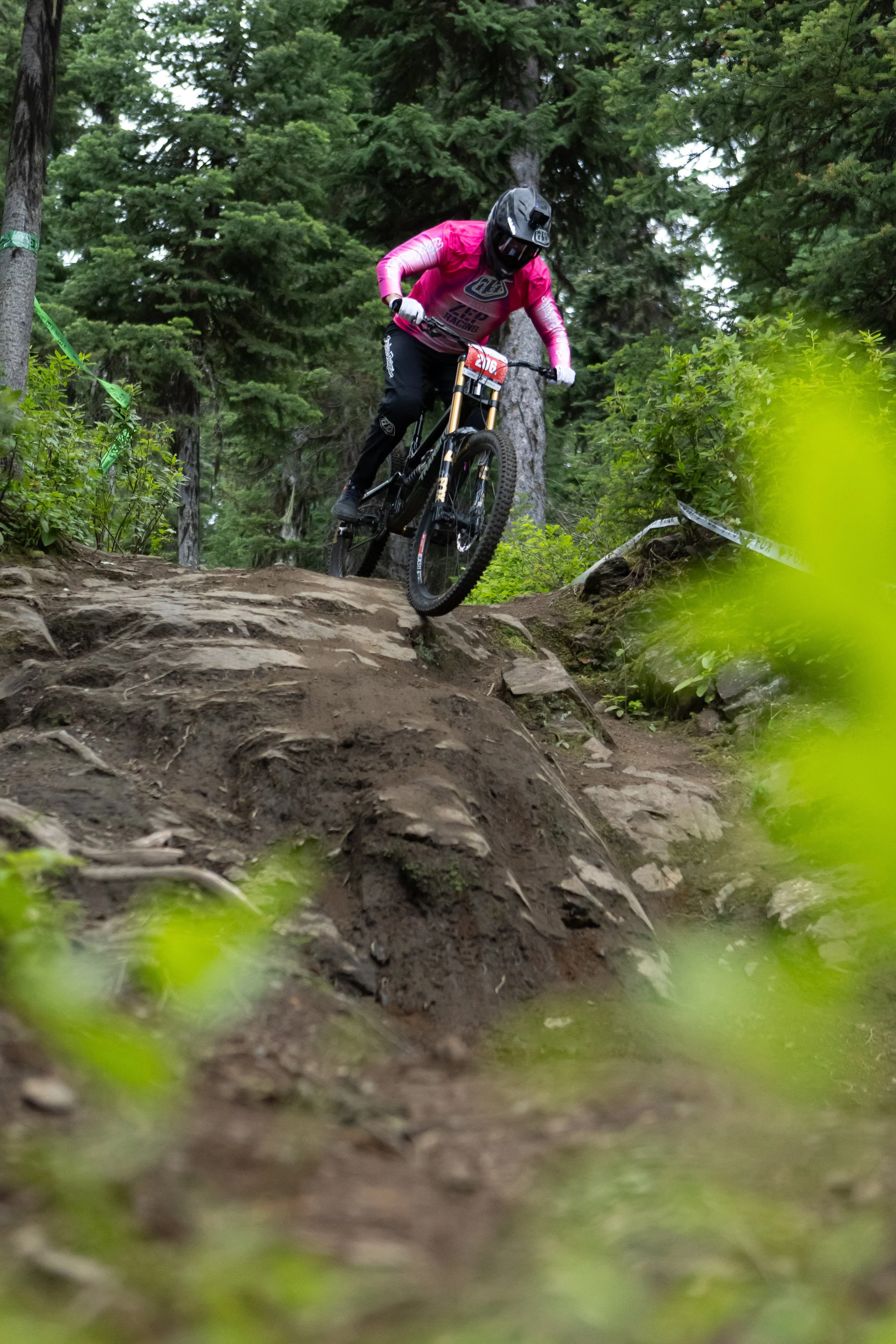 A mountain biker wearing a pink jersey, black helmet, and black pants riding downhill on a dirt trail through a forest with tall green trees.