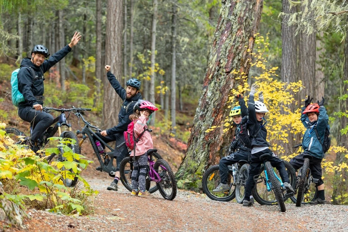 A group of children and adults in helmets and outdoor gear with bikes in a forest, some raising their hands in excitement.