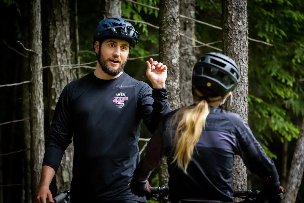 A man in a helmet and black long sleeve shirt talking to a woman in a helmet and black jacket in a forest with trees.