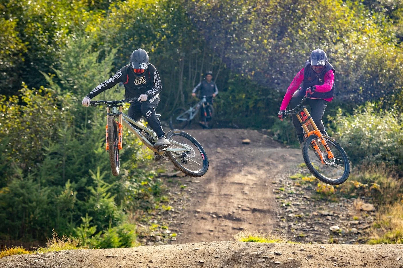 Two mountain bikers in mid-air jumps on a dirt trail surrounded by green trees, with a person on a bicycle in the background.