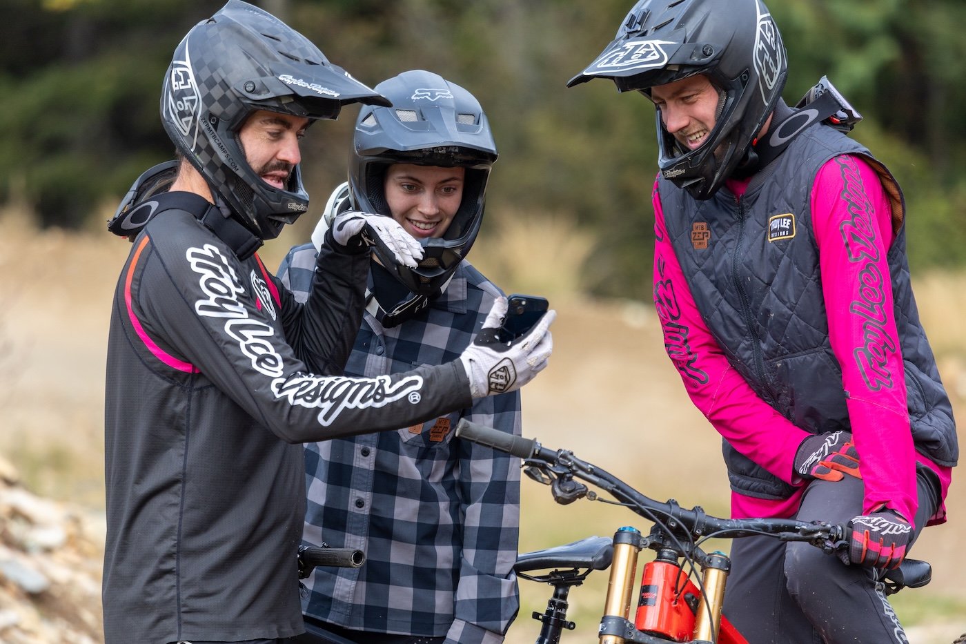 Three mountain bikers wearing helmets and gear looking at a smartphone outdoors.