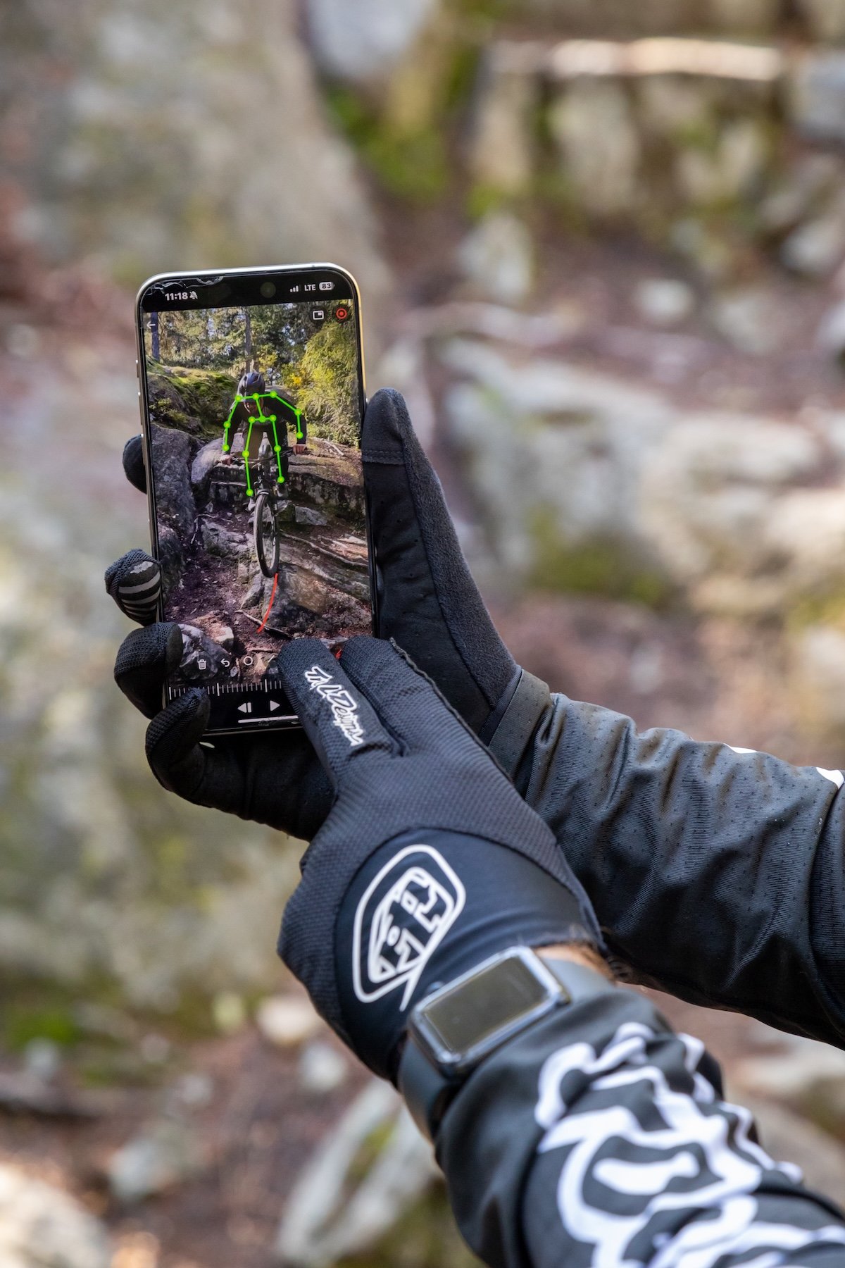 A person wearing black gloves and a black watch holding a smartphone captures a photo of a mountain biker navigating rocky terrain, with the image displayed on the phone including a green skeletal overlay tracking the biker.