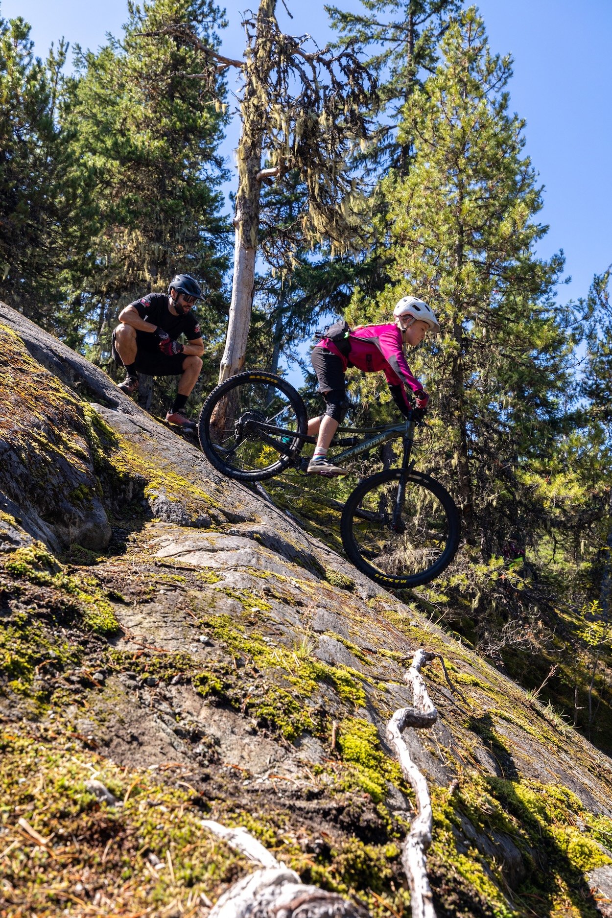 A girl riding a mountain bike down a steep, moss-covered rocky incline in a forest, while a man crouches nearby, observing and guiding her.