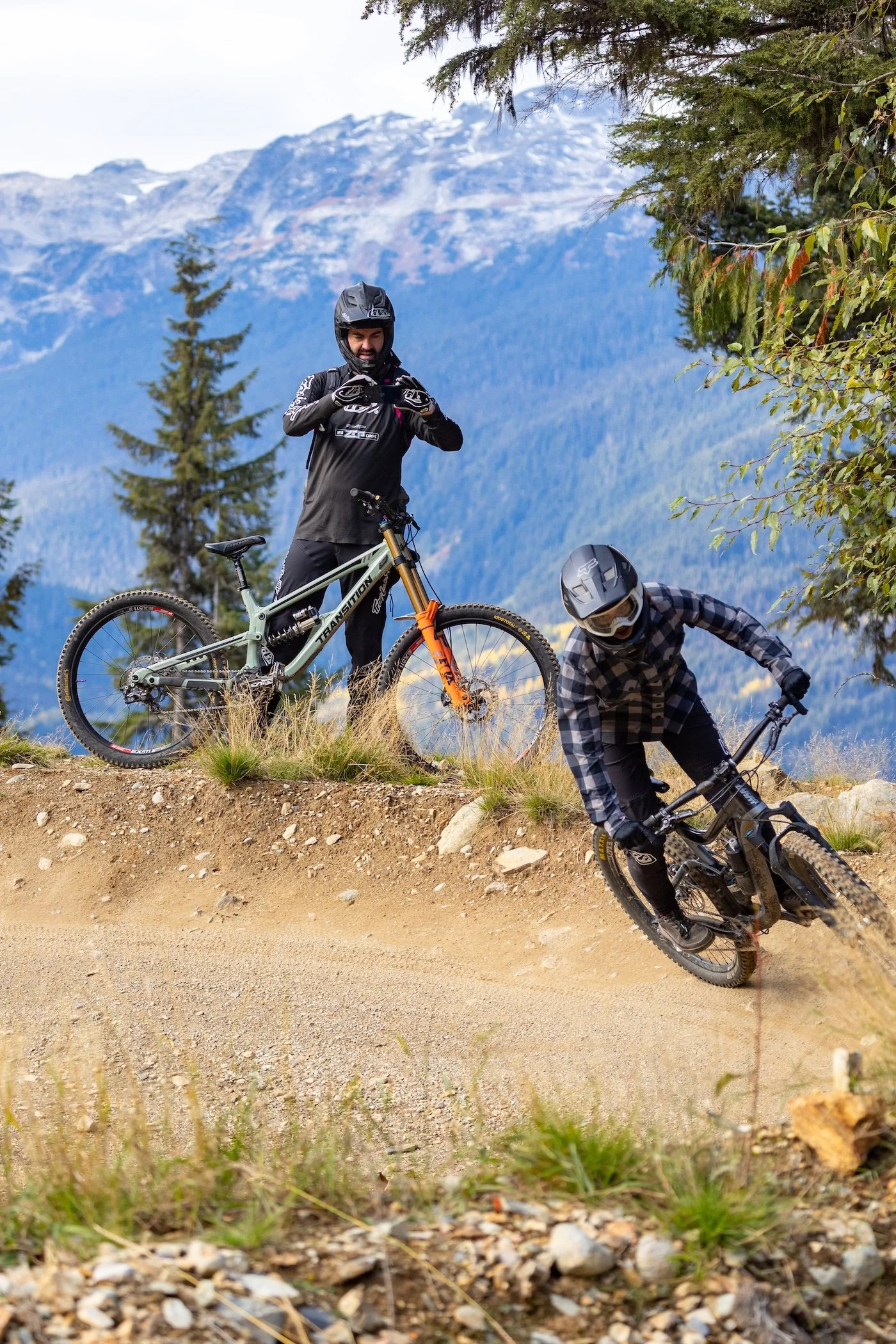 Two mountain bikers on a dirt trail surrounded by trees and mountains in the background. One rider is standing with a bike and the other is leaning into a turn, wearing helmets and outdoor gear.