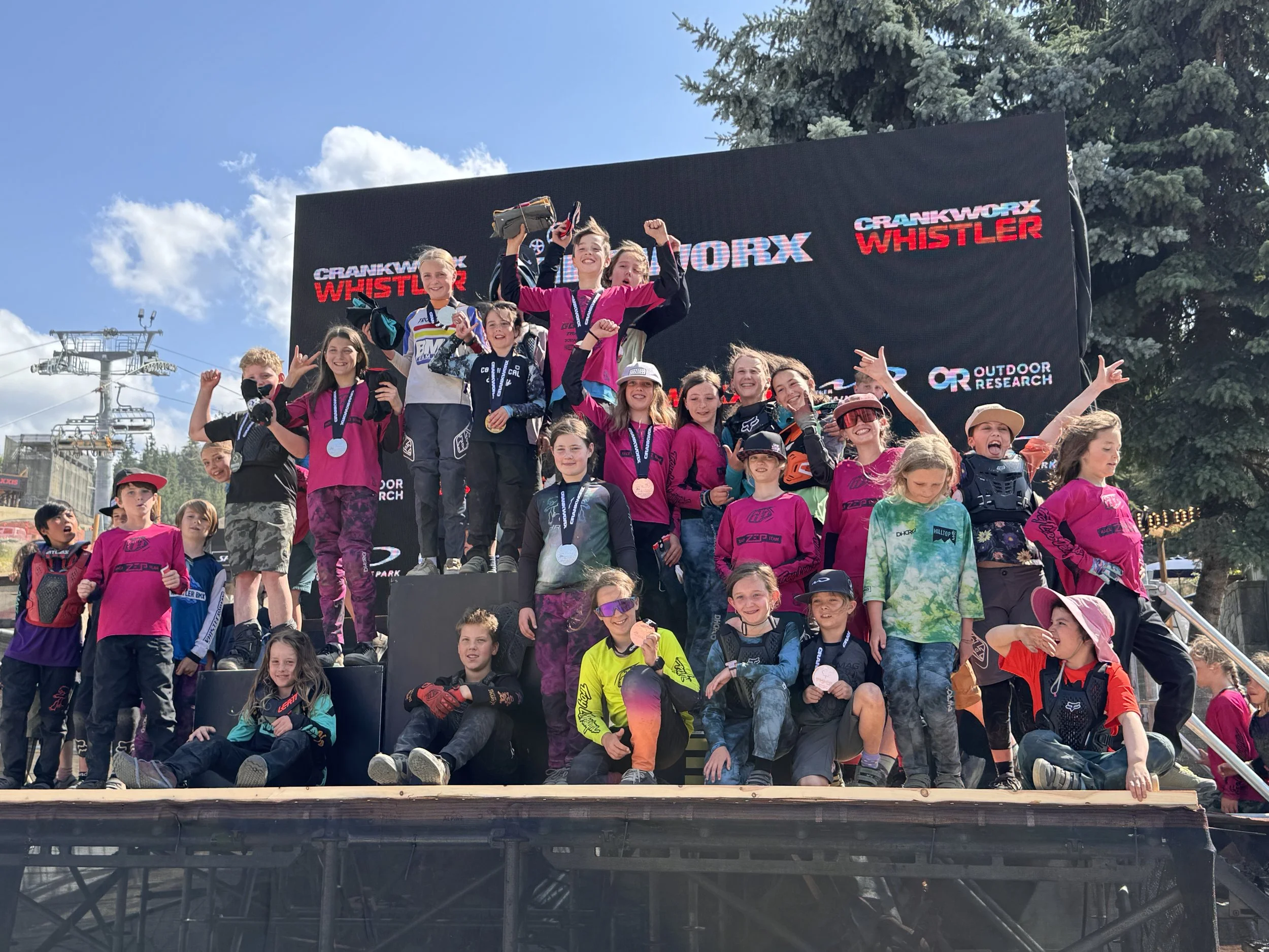Group of children on a podium celebrating with medals after a mountain biking event, with a large digital screen behind them displaying race information and the event name.