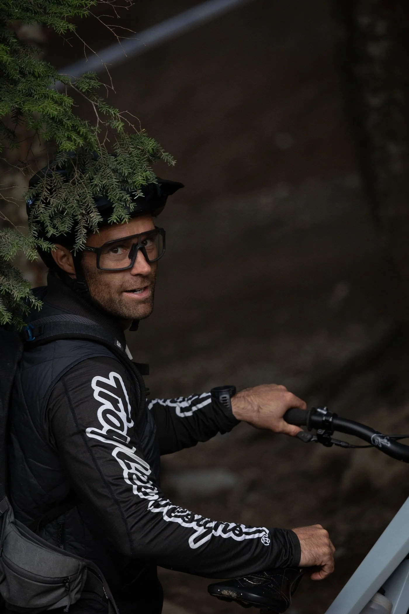 A man wearing a black helmet, glasses, and a black racing jacket with white lettering, riding a mountain bike in a wooded area.