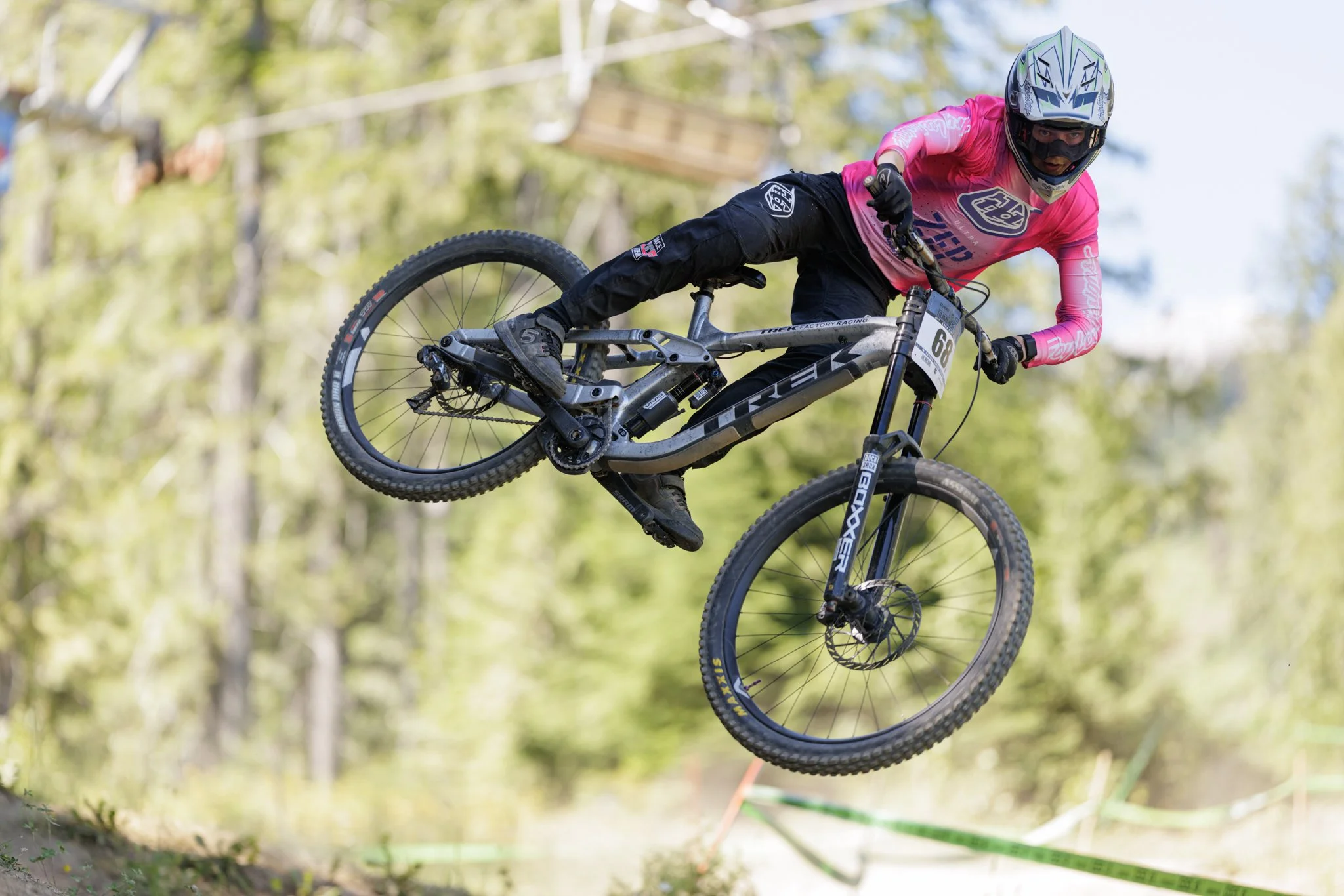 A mountain biker in a pink jersey and black pants is airborne during a jump in a forested trail.
