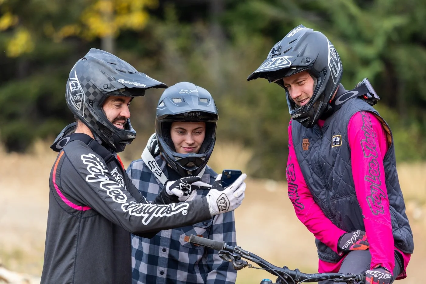 Three off-road mountain bikers wearing helmets and protective gear, looking at a smartphone outdoors in a wooded area.