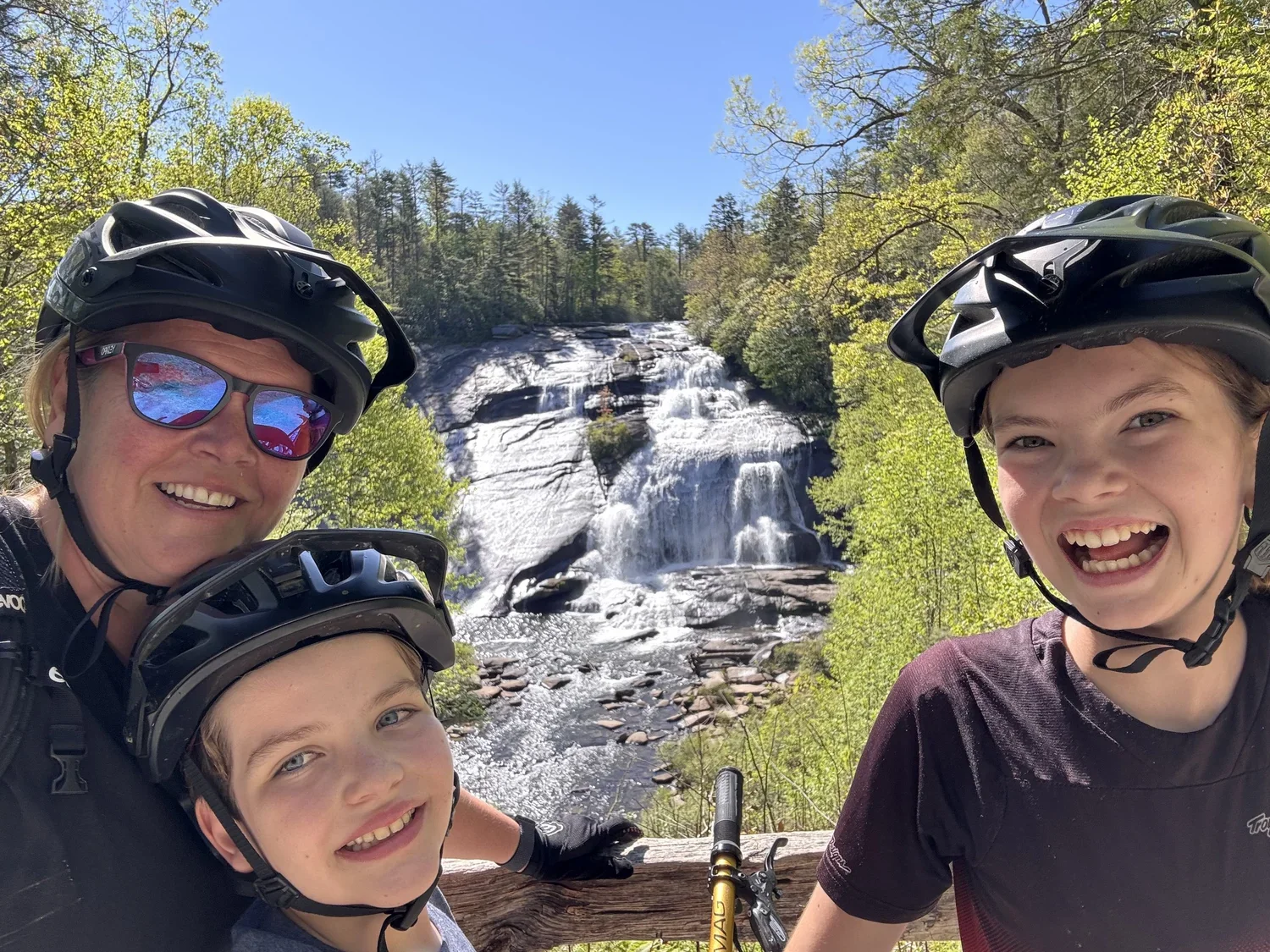 Four people wearing helmets and sunglasses posing for a selfie in front of a waterfall surrounded by green trees on a bright, sunny day.