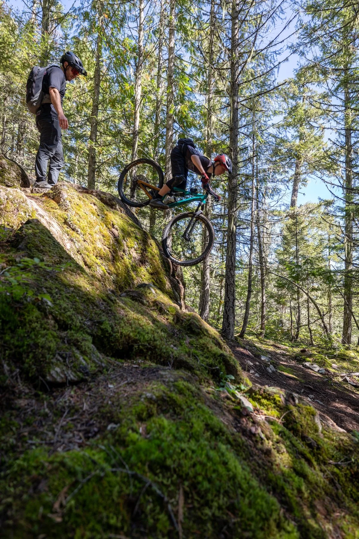 A person mountain biking down a moss-covered rocky slope in a forest with another person watching nearby.