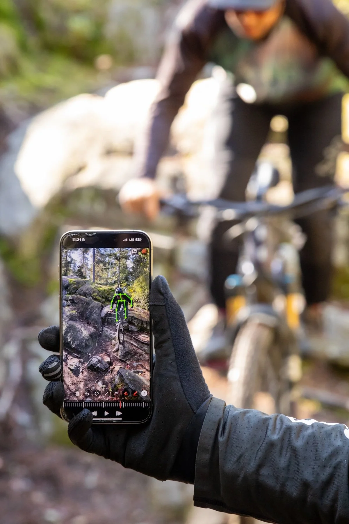 A person holding a smartphone with an augmented reality app that shows a green outline of a person on a mountain bike trail in a forest. The person is wearing gloves and a long-sleeve shirt, with a mountain bike and rocks in the background.