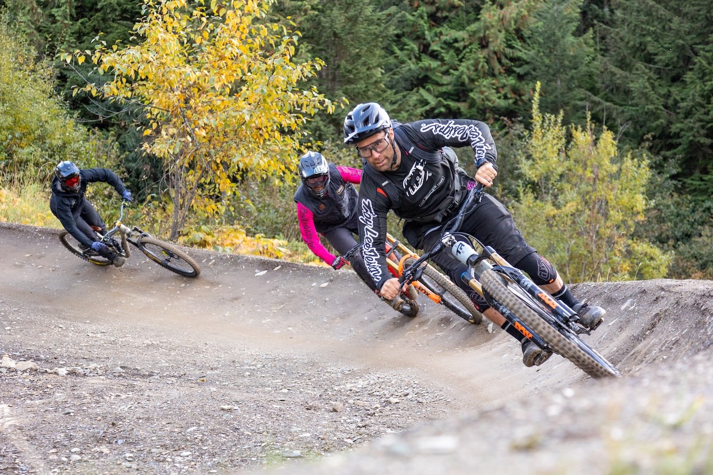 Three mountain bikers wearing helmets and protective gear riding on a dirt trail through a forest with green and yellow trees.