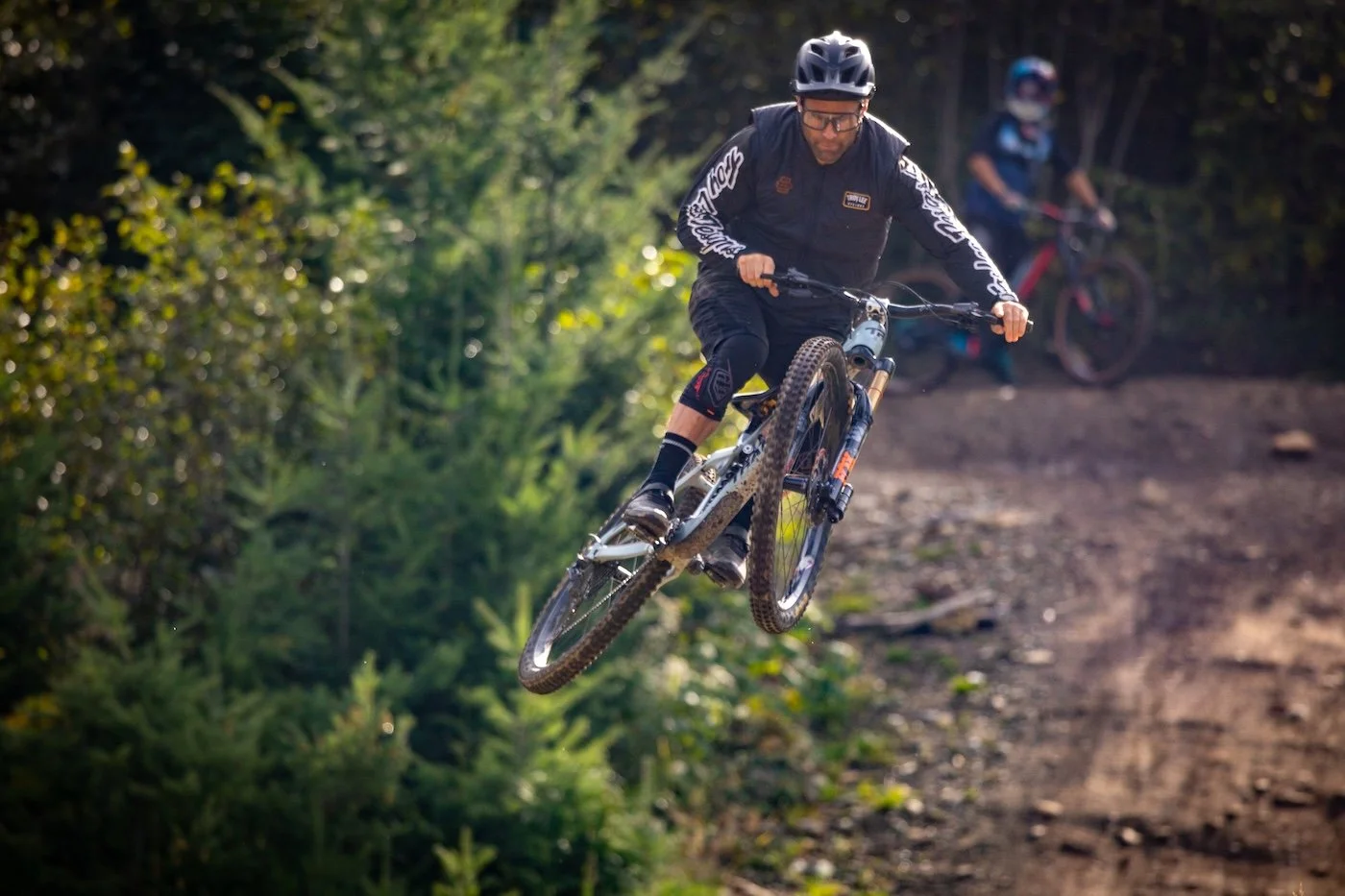 A mountain biker wearing a helmet, black protective gear, and glasses jumps off a dirt trail in a forest.