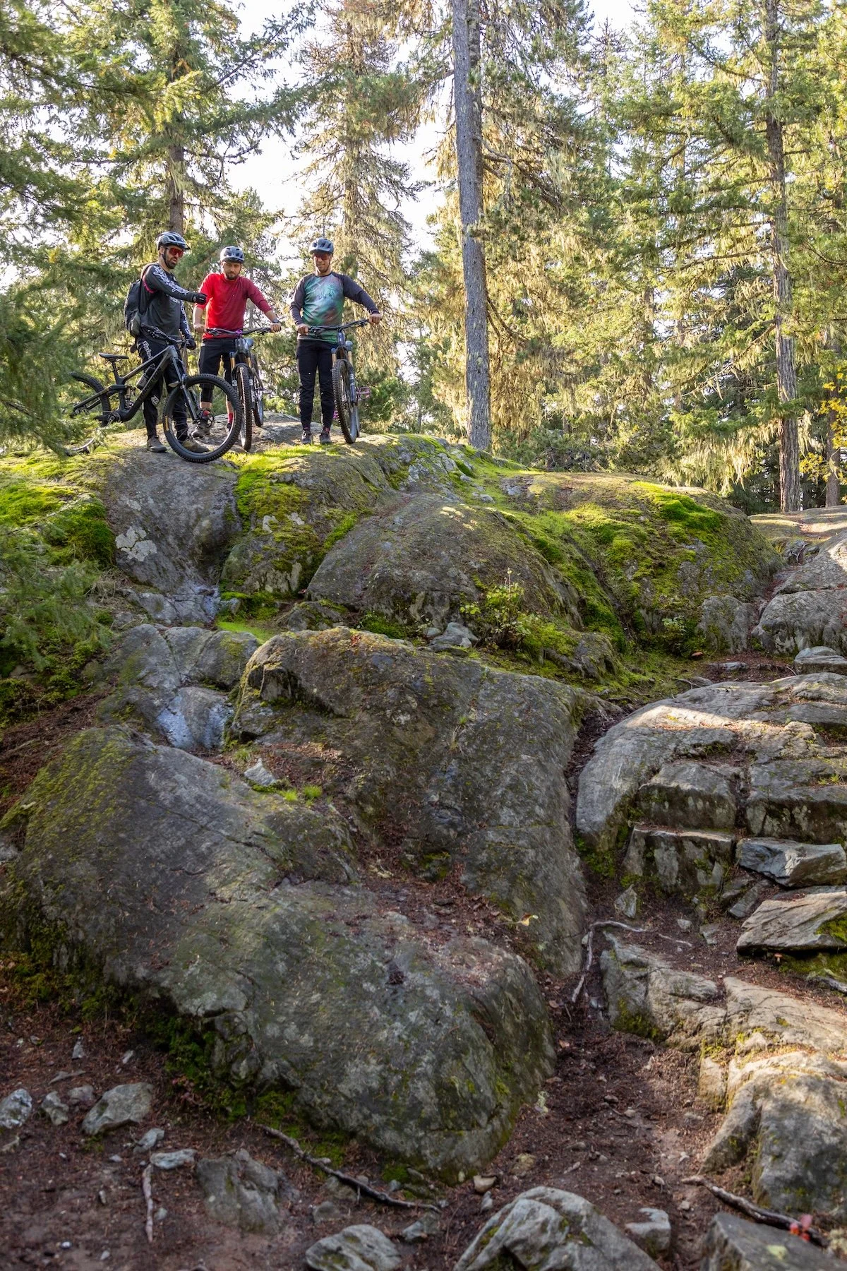 Three men with mountain bikes during a ZEP Tech Master Clinic standing on mossy rocks in a forested area, wearing helmets and outdoor gear.