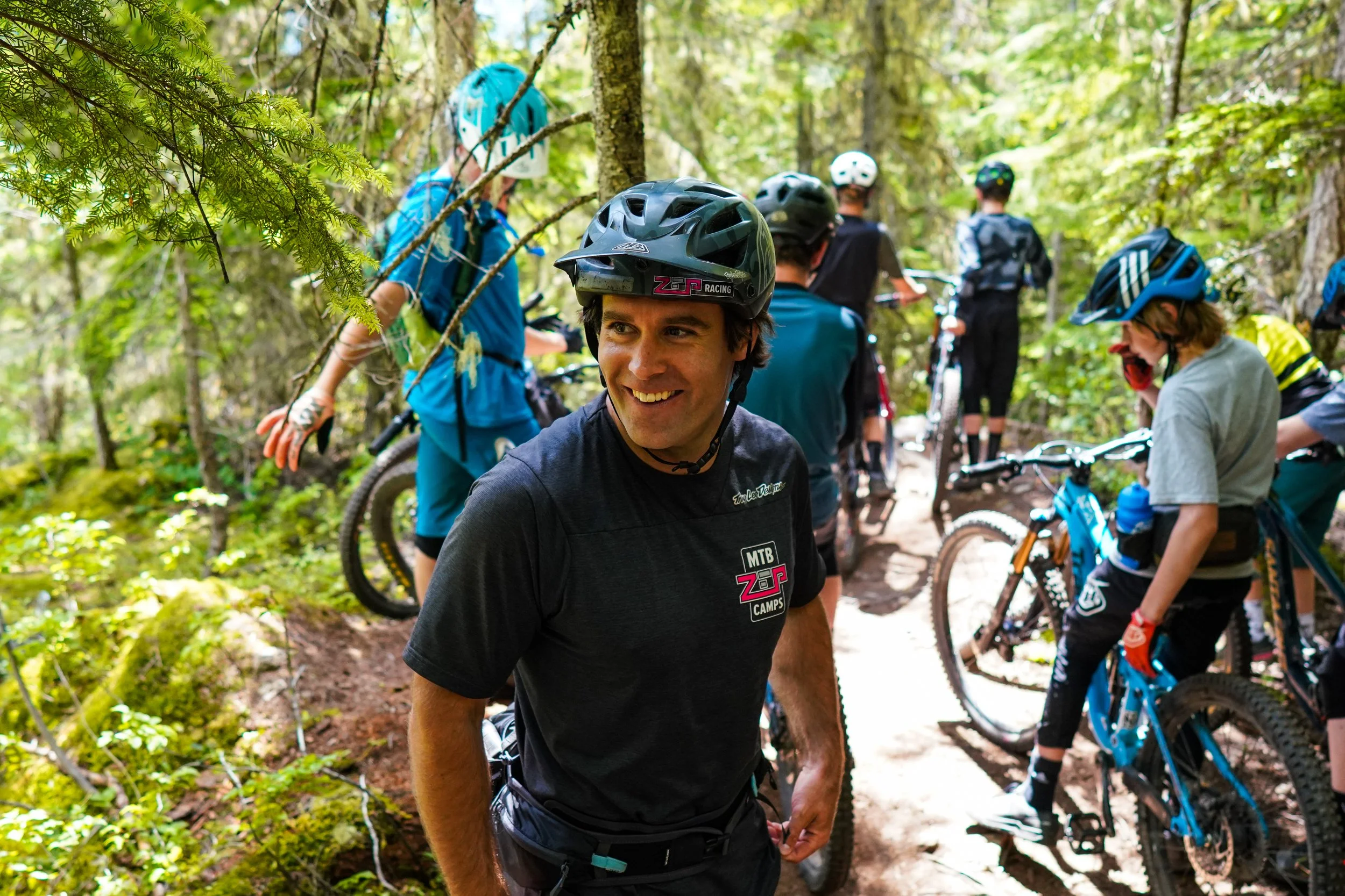 A group of mountain bikers taking a break on a forest trail, with one smiling man in the foreground wearing a black bike helmet and black MTB camp shirt.