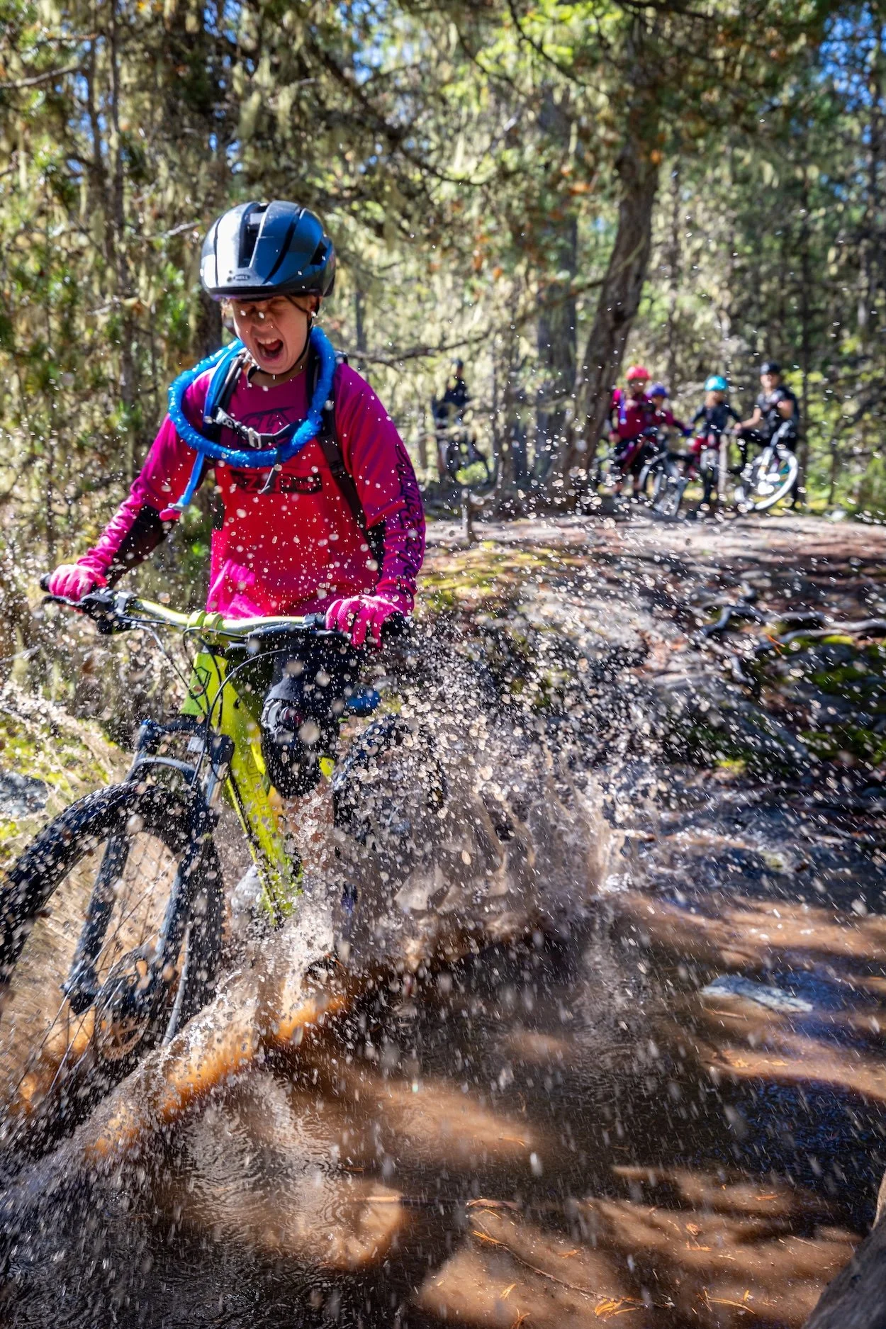 A young girl wearing a black helmet and pink gloves rides a bicycle through a muddy trail in a forest, splashing water. In the background, other children with helmets and bikes are visible on the trail.