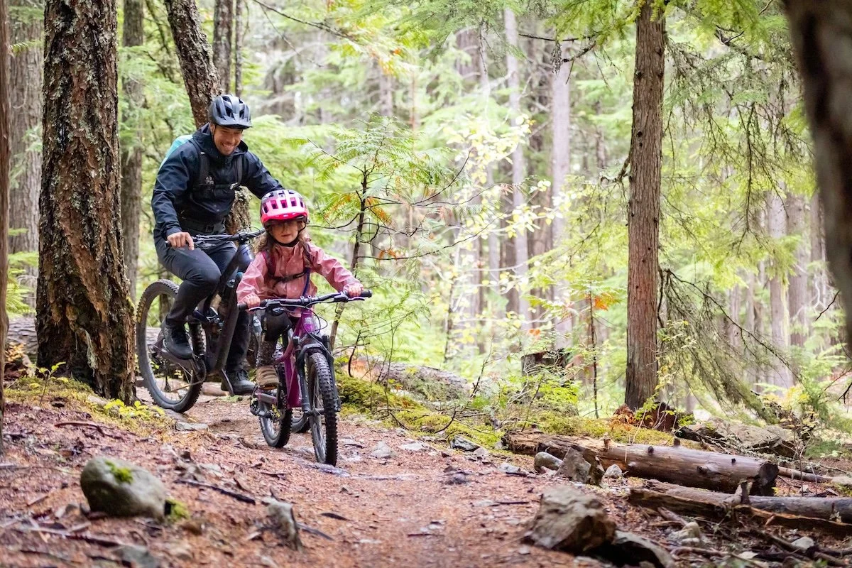 A man and a young girl riding mountain bikes along a dirt trail in a forest, both wearing helmets.