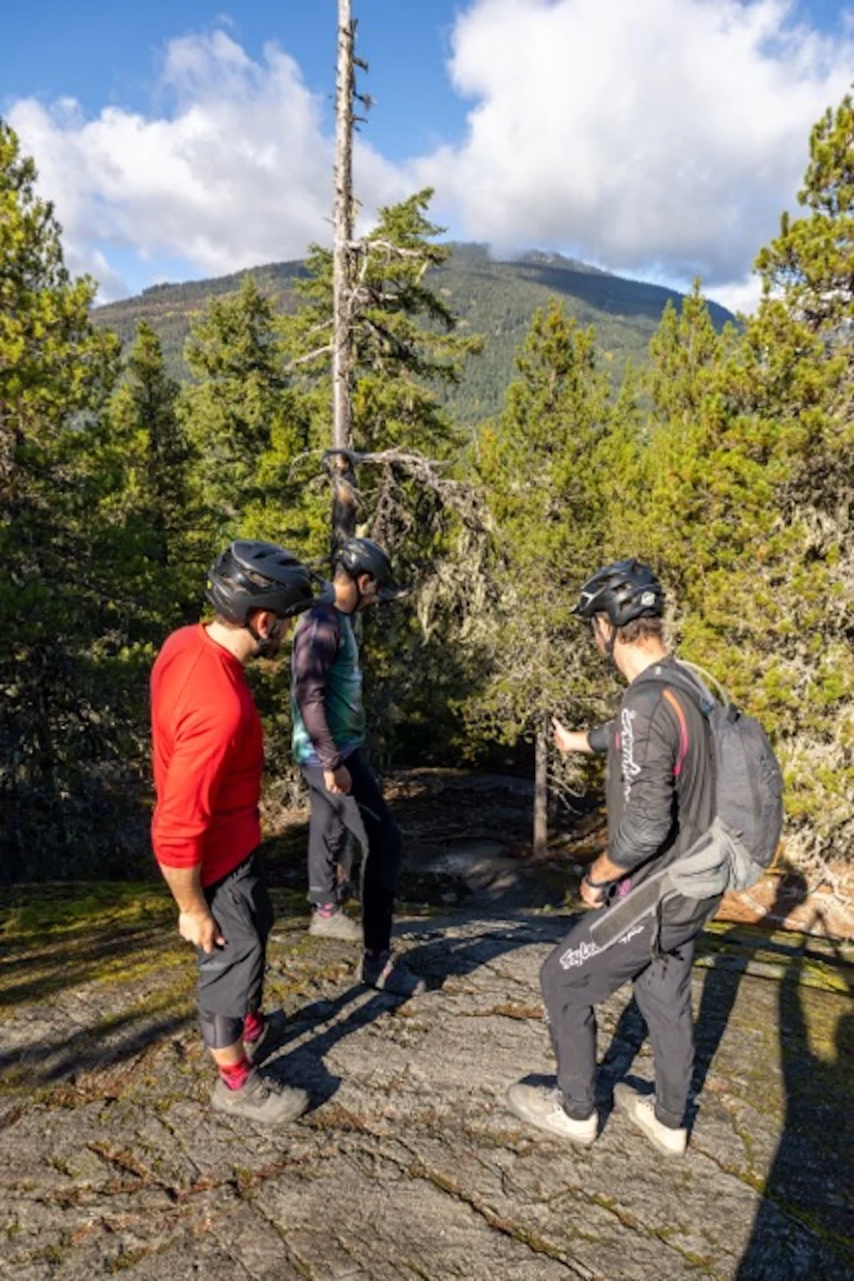 Three people wearing helmets and outdoor gear standing on a rocky trail in a forested mountainous area, with trees, a mountain in the background, and partly cloudy skies.