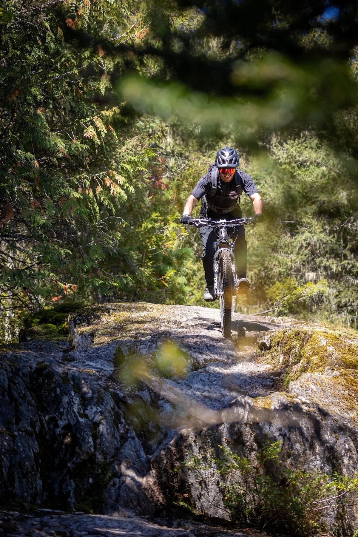 A person mountain biking on a rocky trail surrounded by dense forest with trees and green foliage.
