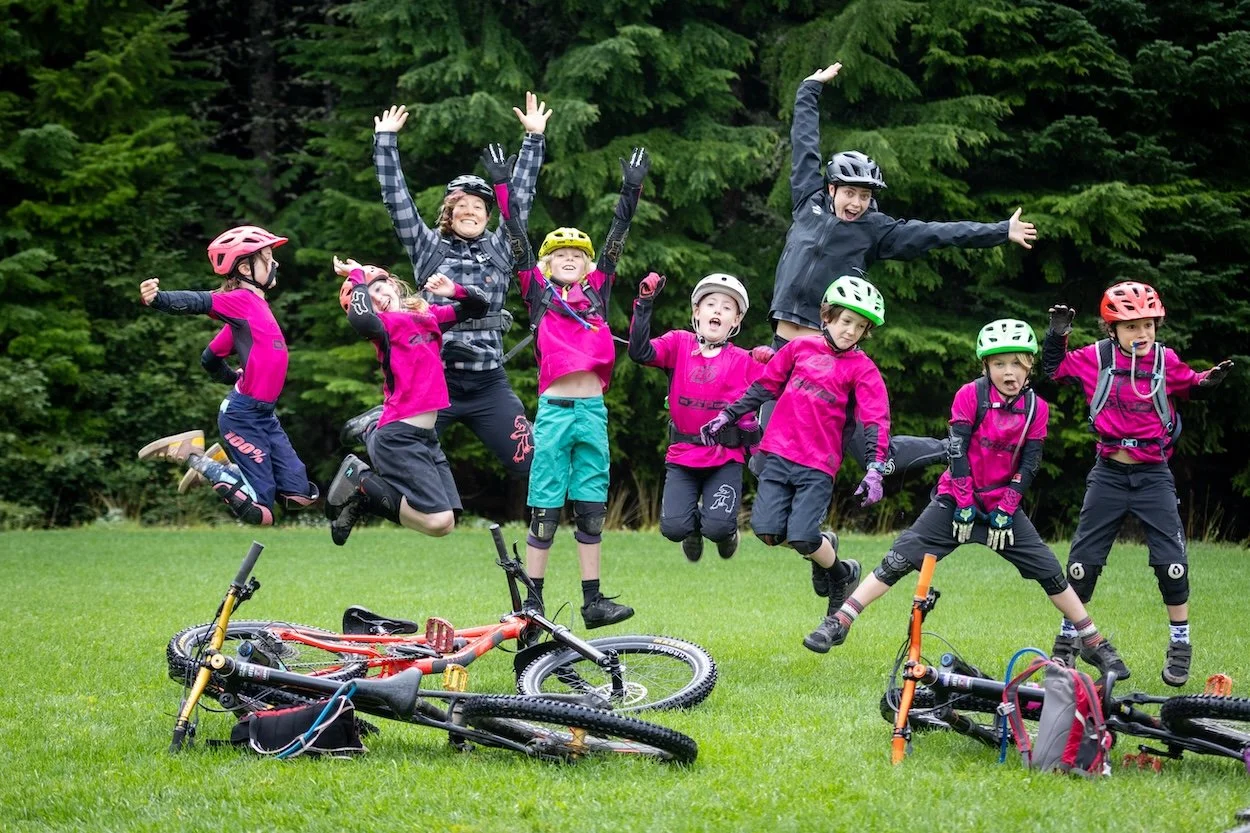 Group of children and two adults in helmets jumping in the air on a grassy field, with mountain bikes lying on the ground, surrounded by trees.