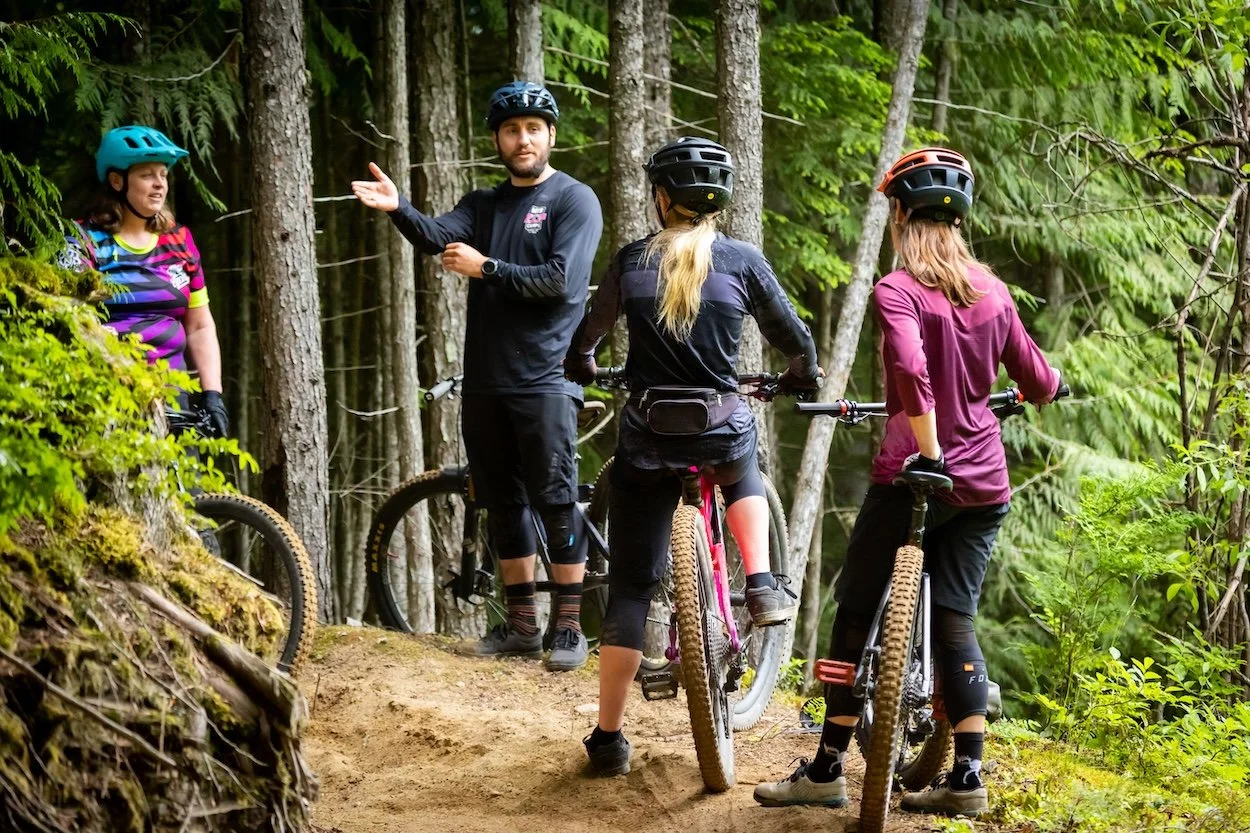 A group of four mountain bikers, three women and one man, are on a trail in a dense forest, with the man giving instructions while the women listen and hold their bikes.