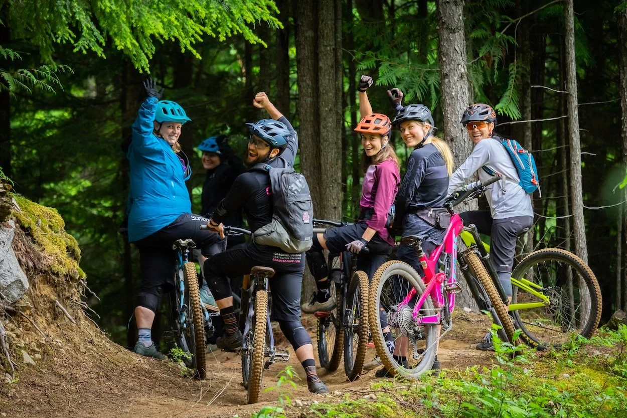 Group of five women mountain biking in a forest, wearing helmets and gear, smiling and enjoying the outdoor adventure.