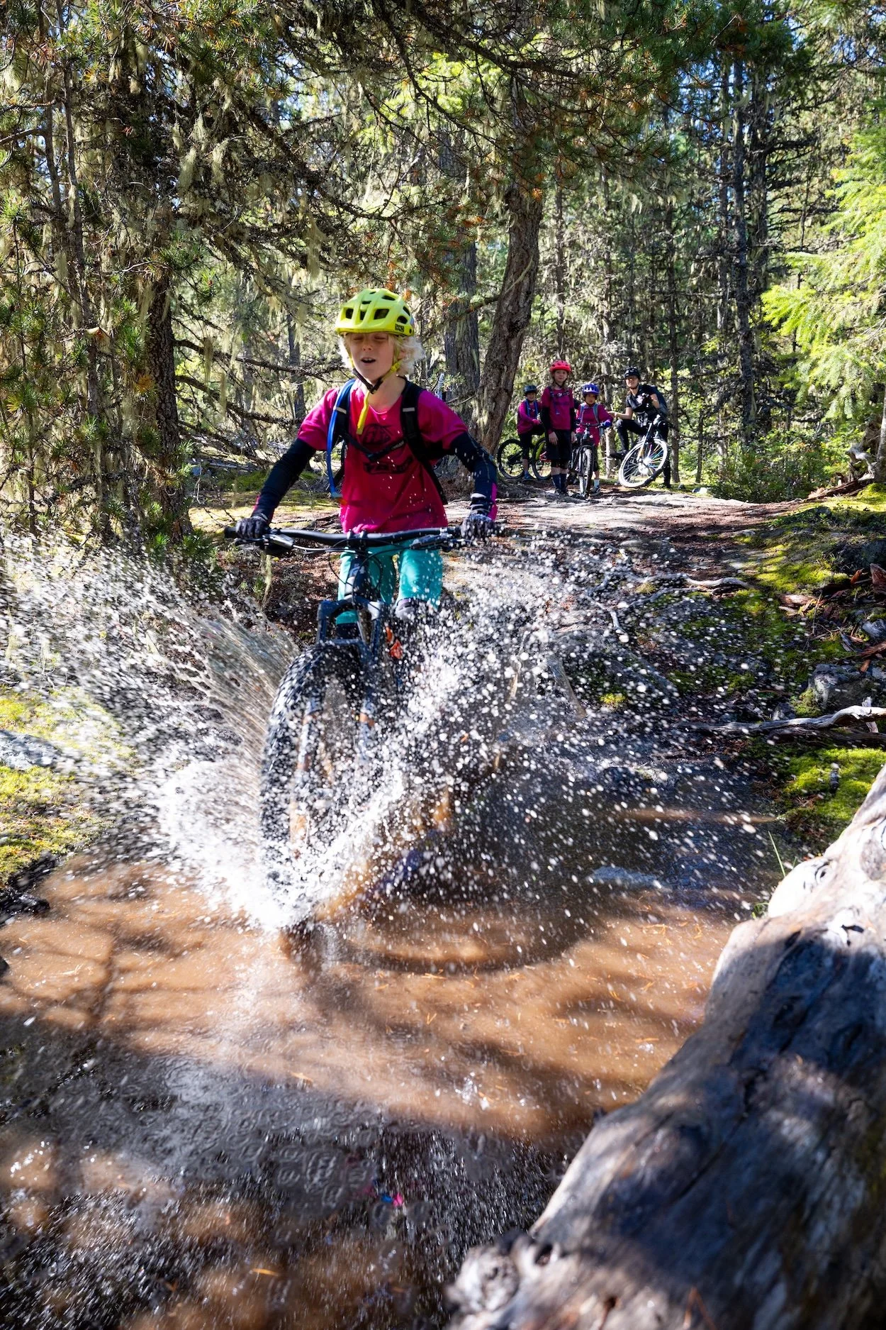 A young girl wearing a yellow helmet and pink shirt rides a bike through a shallow stream in a forest, splashing water as she crosses. Four people on bikes follow behind her on a trail surrounded by trees.