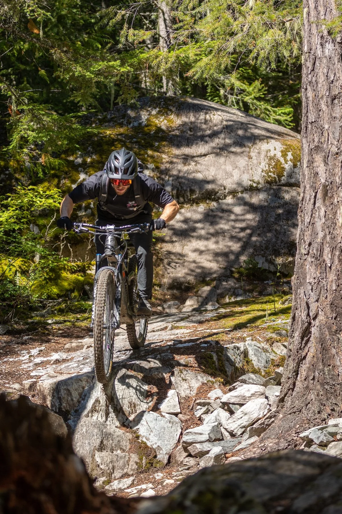 A mountain biker in black gear, wearing a helmet and sunglasses, riding downhill on a rocky trail in a forest.