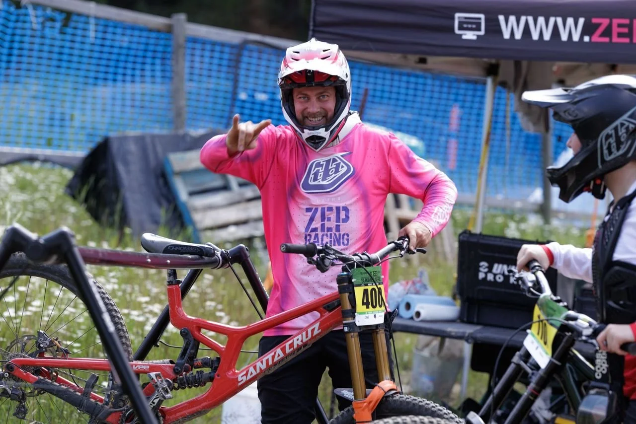 A man in a pink mountain biking jersey and helmet making a hand gesture while standing next to a red Santa Cruz mountain bike at an outdoor bike park.