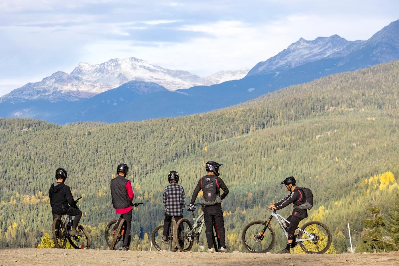 Five mountain bikers wearing helmets and gear, with some carrying backpacks, standing on a dirt trail overlooking a lush green forest with snow-capped mountains in the background.
