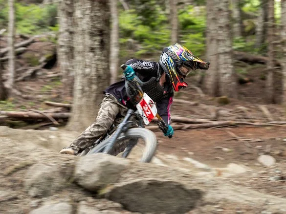 A person mountain biking downhill through a forest, wearing a helmet, goggles, and protective gear.