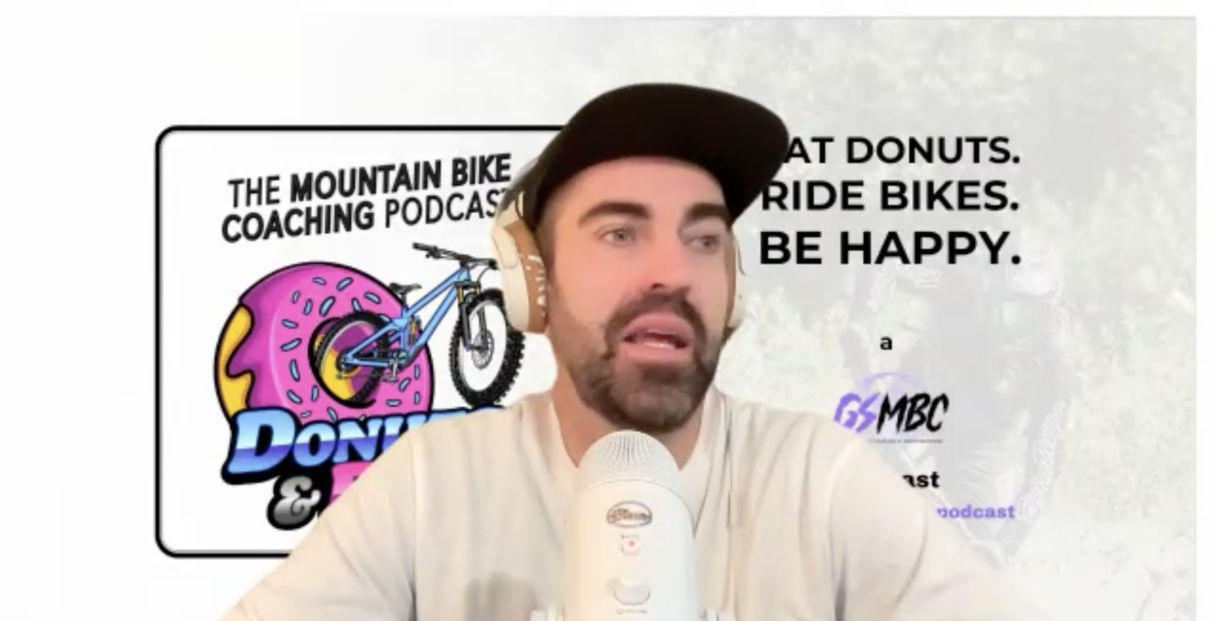 A man with a beard and mustache wearing a black baseball cap and beige shirt, speaking into a microphone with a background poster. The poster shows a colorful illustration of a bike riding through a pink donut with yellow icing, and contains the text 'The Mountain Bike Coaching Podcast' and phrases about donuts, riding bikes, and happiness, along with the logo 'GS MBC'.