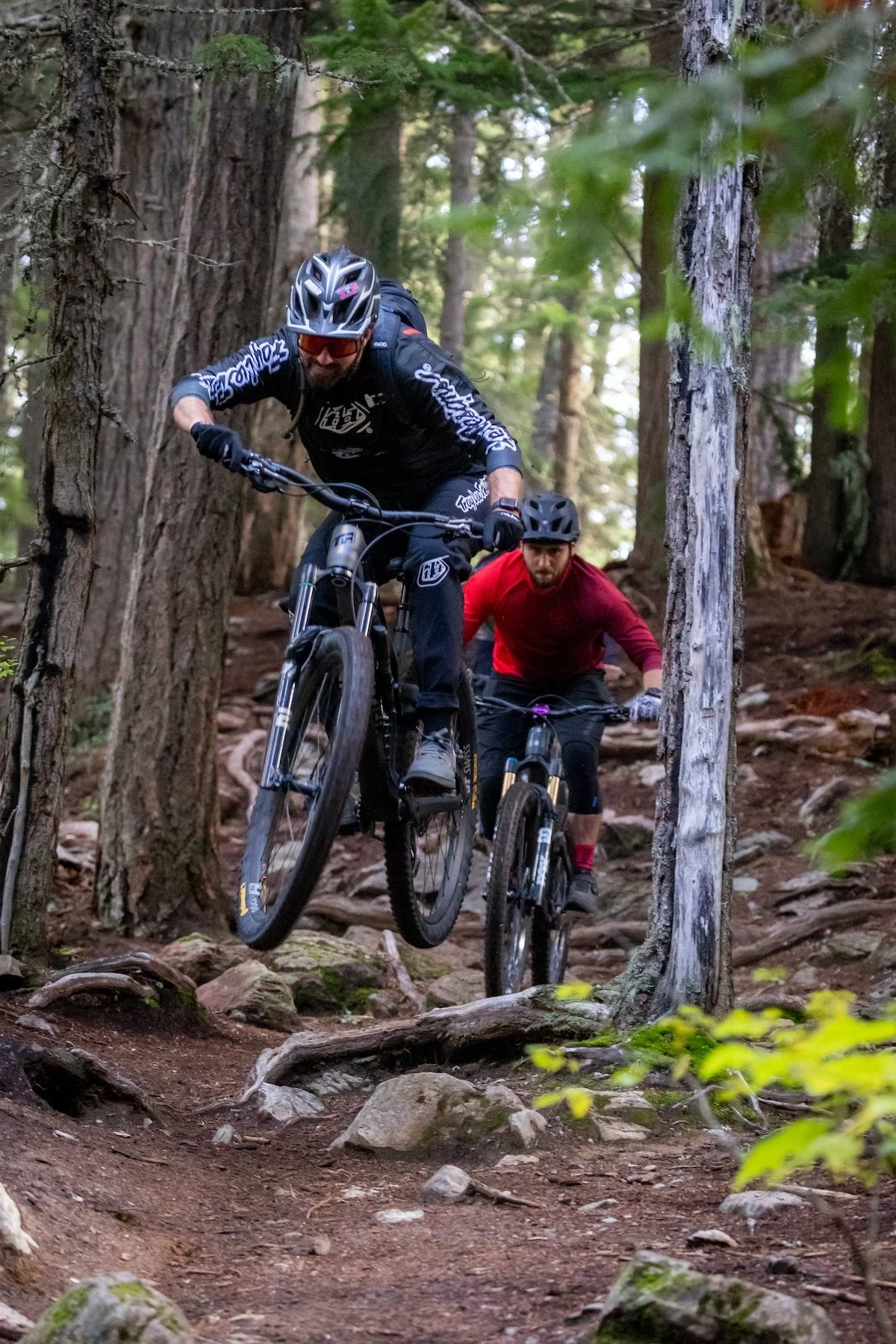 Two mountain bikers riding on a rocky trail in a dense forest. The rider in front is airborne, wearing a helmet with goggles and a black outfit, while the other rider follows behind in a red shirt and helmet.