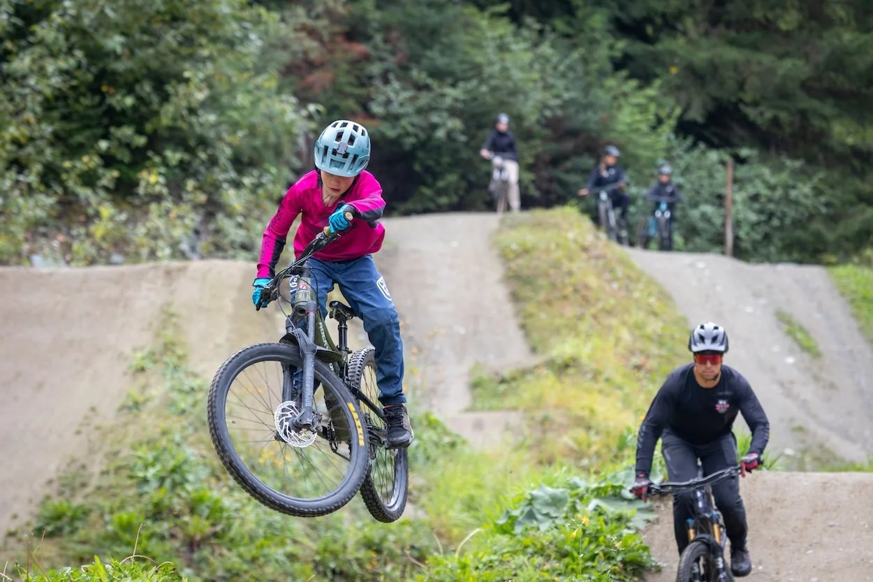Children riding bikes on dirt jumps in a wooded area, with one child airborne and others biking on the ground.