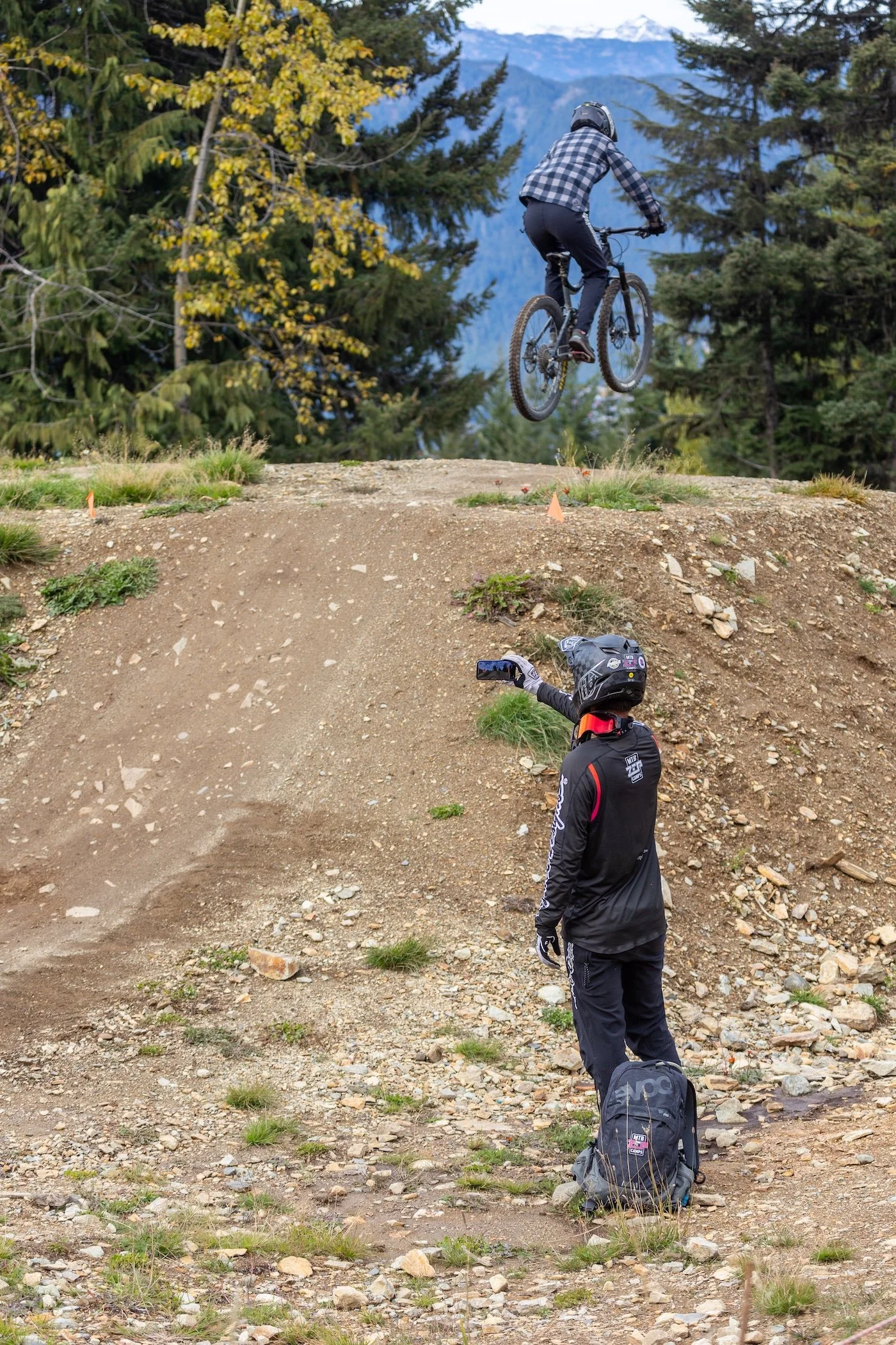 A mountain biker in mid-air jump during a ZEP Bike Park lesson, on a dirt trail in Whistler Bike Park with a ZEP coach watching and recording with a smartphone. The scene is set in a forest with mountains in the background.