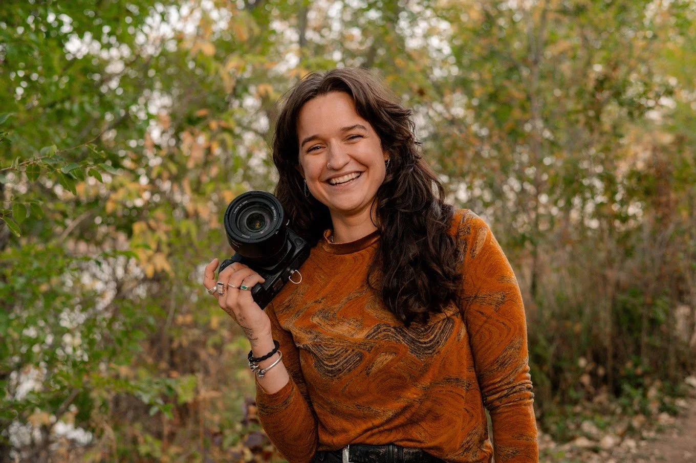 A woman with long dark hair smiling while holding a camera outdoors in a wooded area with autumn leaves.