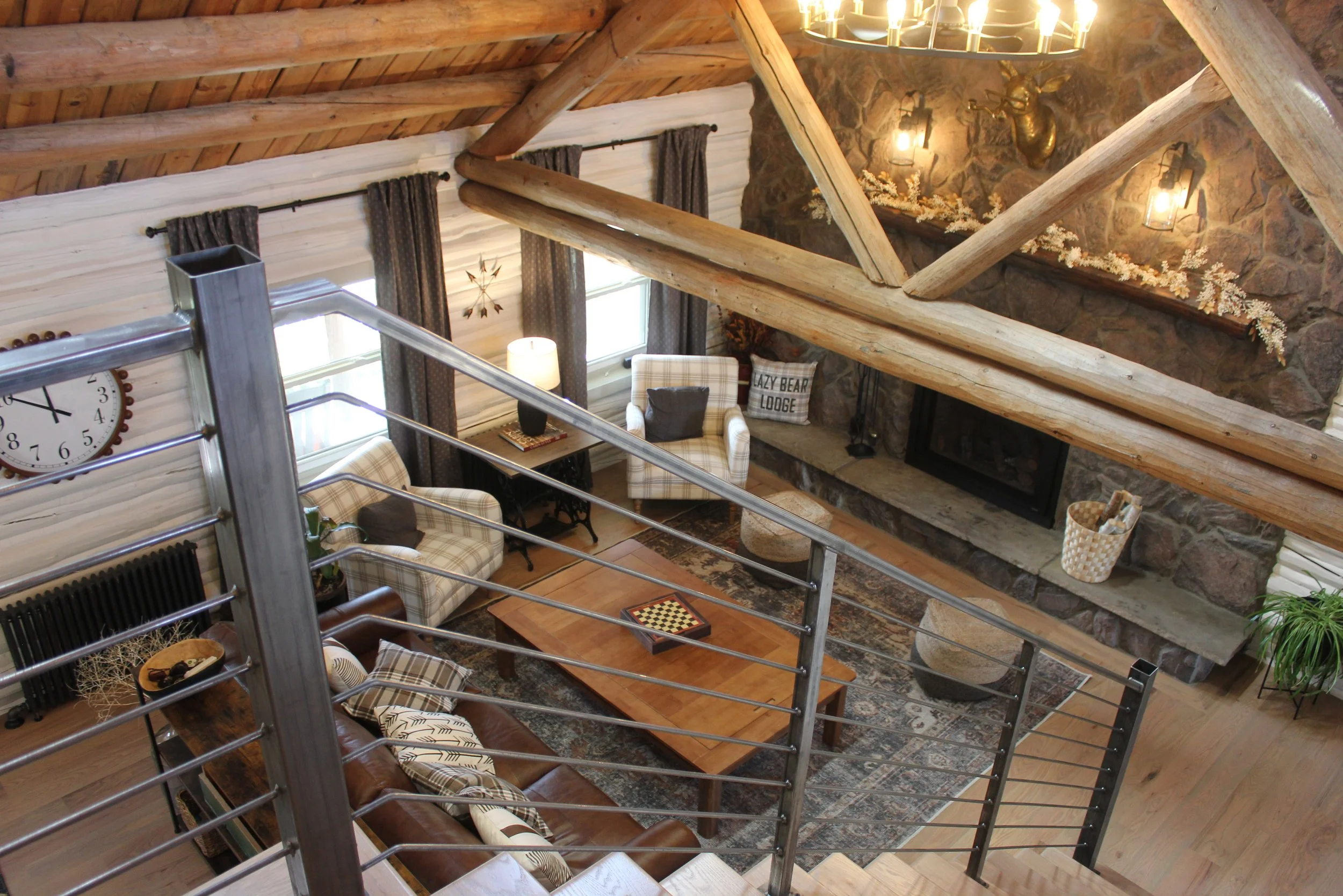 Interior view of a rustic living room with a stone fireplace, plaid armchairs, a wooden coffee table, and log beams on the ceiling, seen from above.